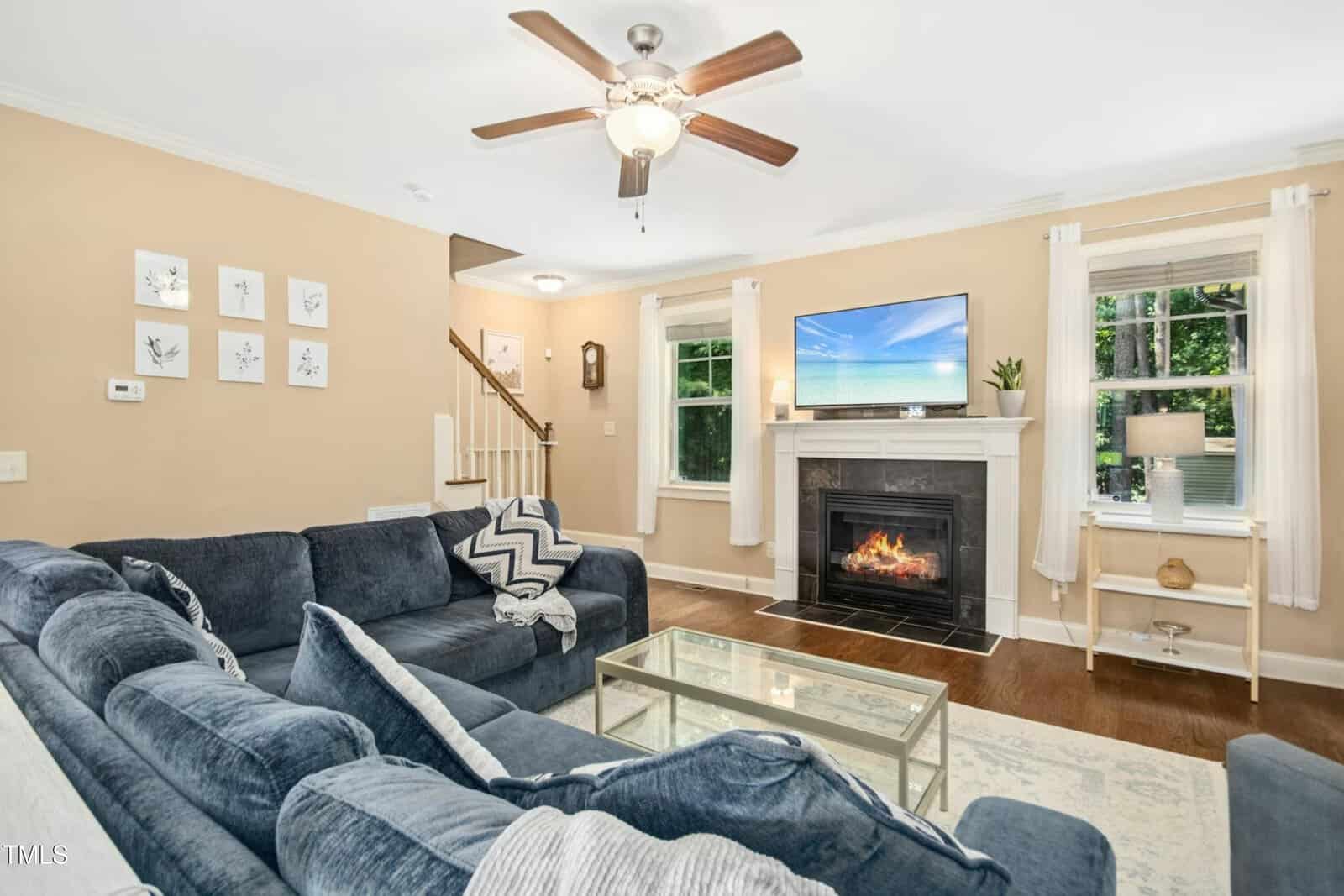 A cozy living room with a blue sectional sofa, a glass coffee table, and a fireplace with a TV above it. The walls are beige, and two windows with white curtains let in natural light. A ceiling fan hangs above.