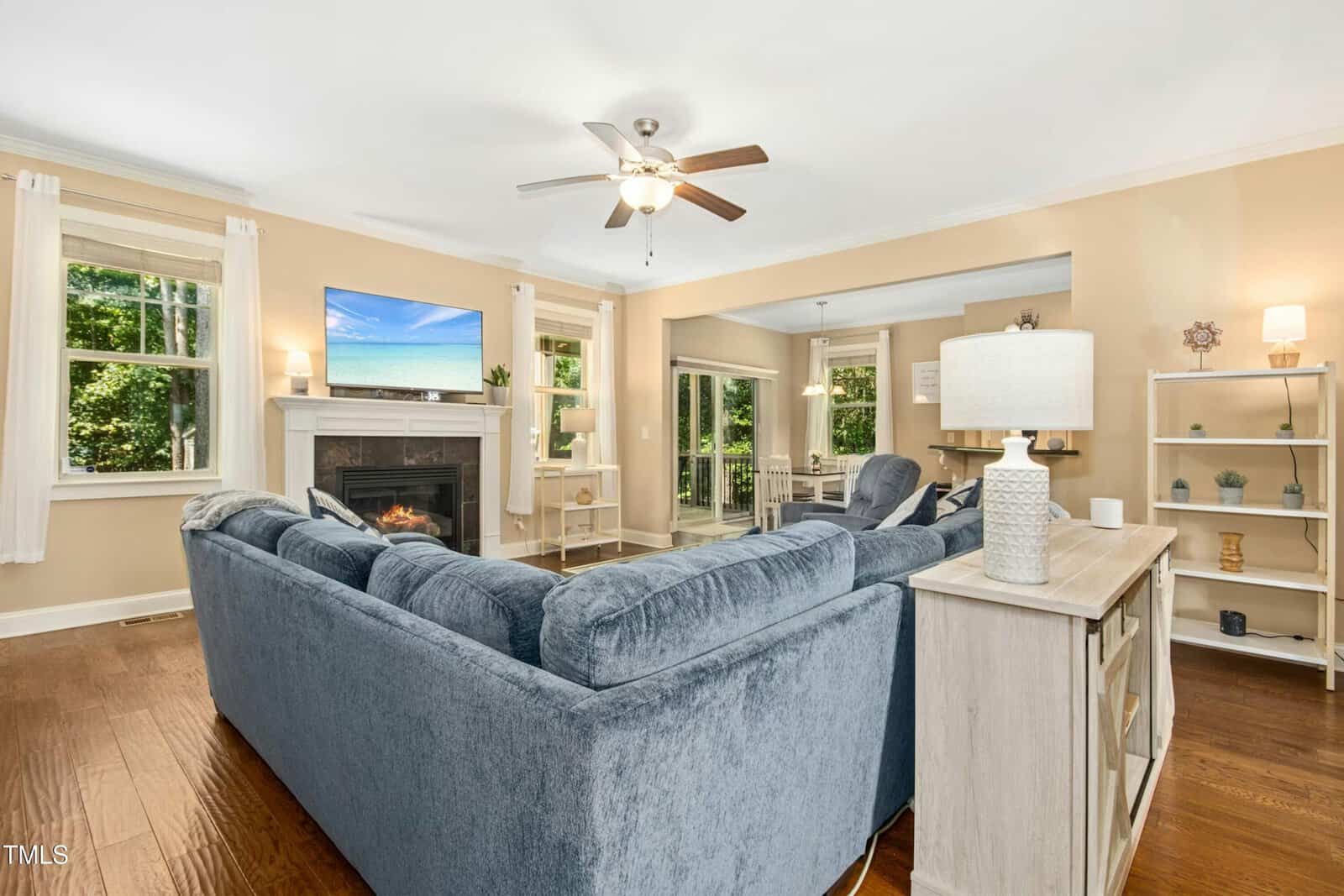 A living room with a blue sectional sofa, a fireplace, a mounted TV, ceiling fan, shelves with decor, and large windows. Light walls and wood flooring. Dining area and deck visible through openings.