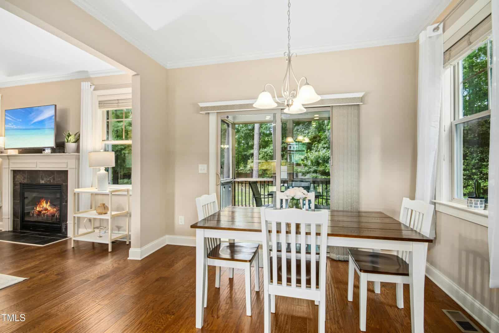 Dining area with a wooden table and four white chairs, a window with white curtains, glass doors leading to a deck, hardwood floors, and partial view of a living room with fireplace and TV.