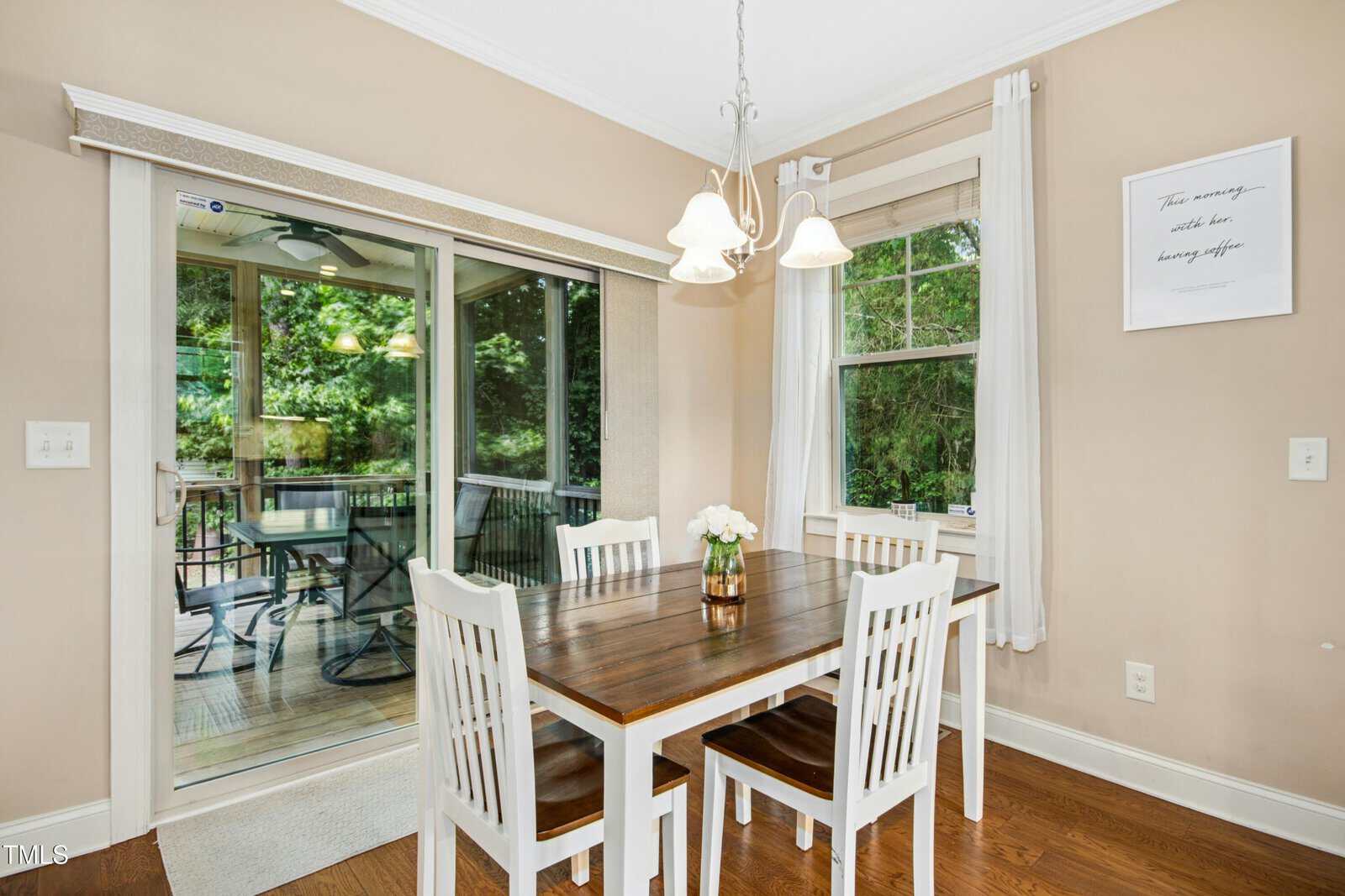 A dining area with a wooden table, four white chairs, a small flower vase, two windows with white curtains, hanging light fixture, and a sliding glass door leading to a screened porch with outdoor seating.
