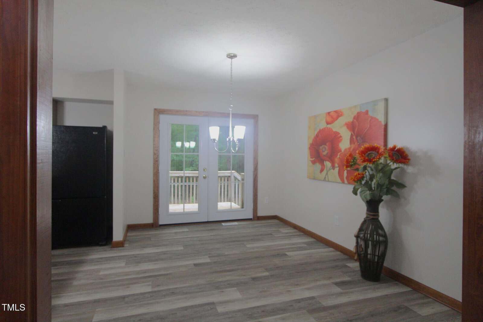 Bright dining area with wooden floors, a chandelier, double glass doors to a deck, a large vase with orange flowers, a matching floral painting, and a glimpse of a black refrigerator in the adjacent kitchen.