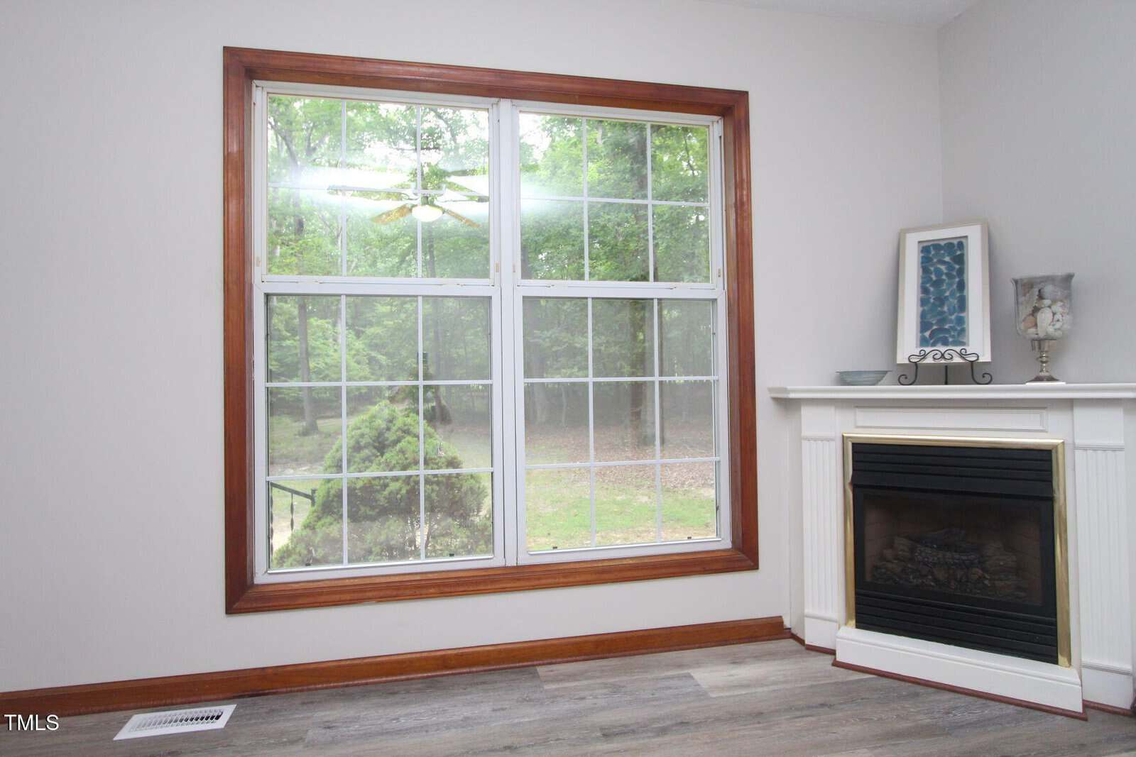 Large window with wooden trim overlooks a green, wooded yard; a white fireplace with a framed picture, candles, and a dish sits on the right, atop gray wood flooring.