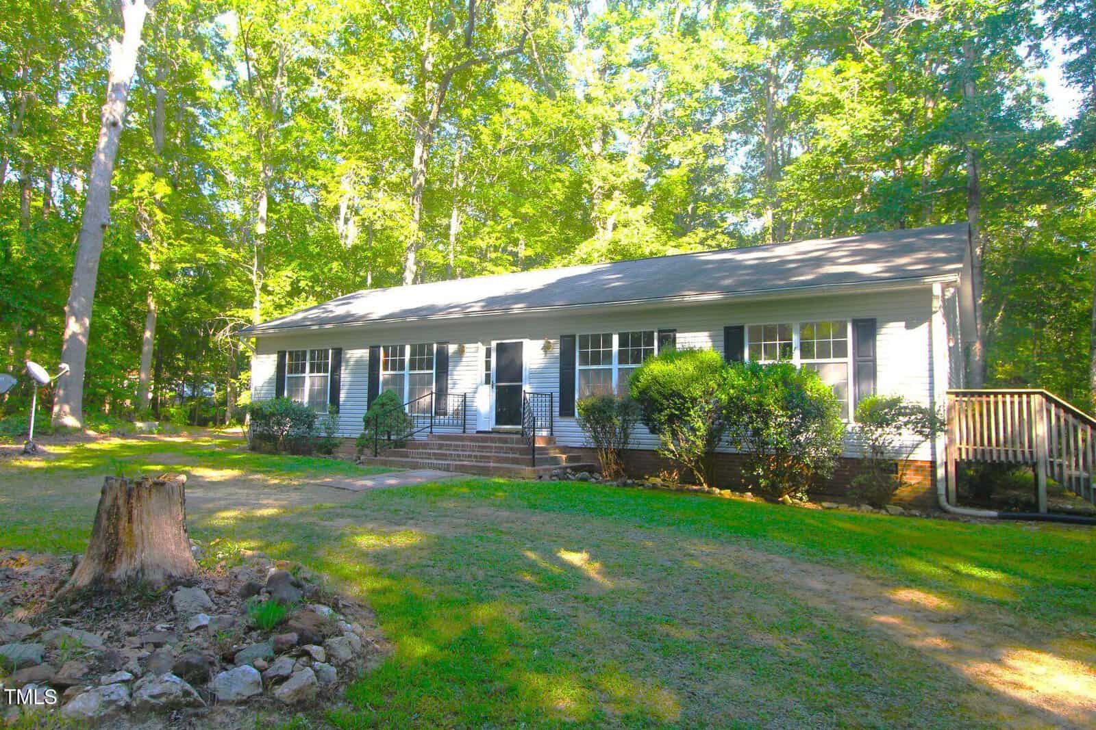 Charming single-story house at 622 Enon Road, Oxford, with light-colored siding, black shutters, and a dark roof. Enjoy a front porch with railings, bushes along the front, and a tree stump in the grassy yard surrounded by trees.