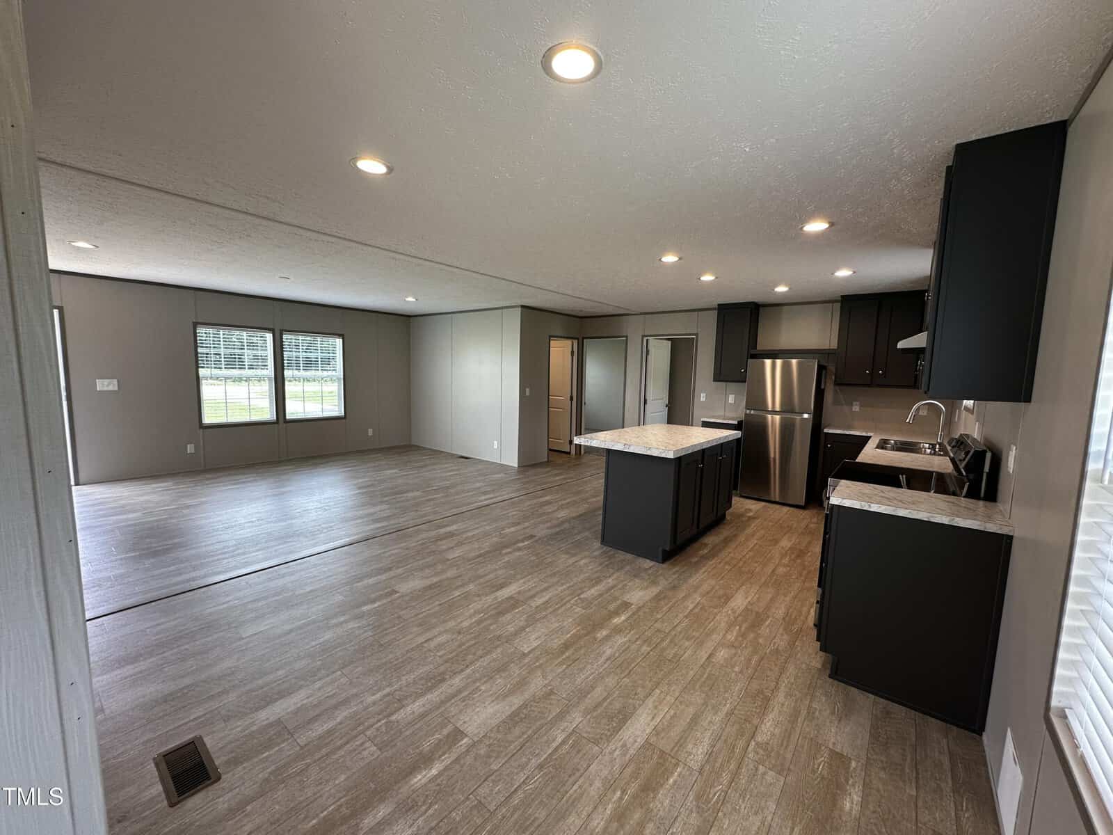 Open-concept living space with wood-look flooring, recessed lighting, large windows, and a kitchen featuring dark cabinets, stainless steel appliances, and a central island. Light-colored walls and ceiling throughout.