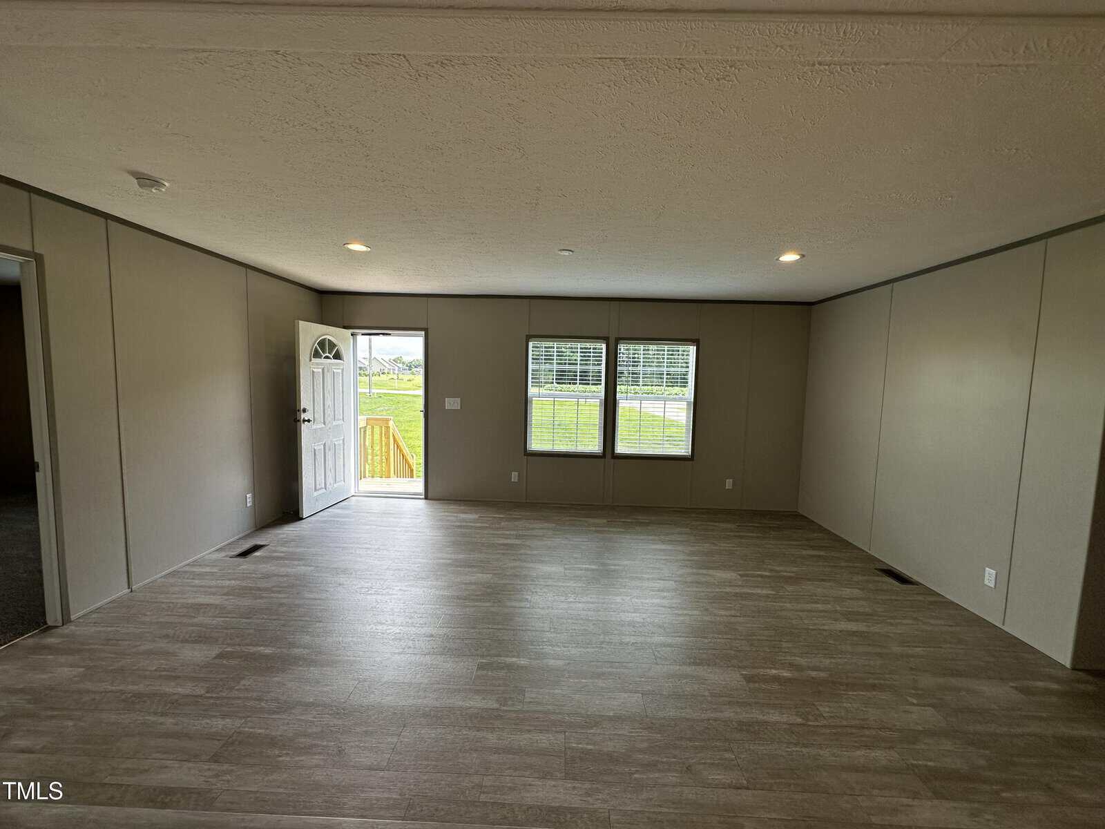 Spacious empty room with wood-look flooring, beige walls, and two large windows letting in natural light. An open white door leads outside to a grassy yard. Ceiling lights are evenly spaced.