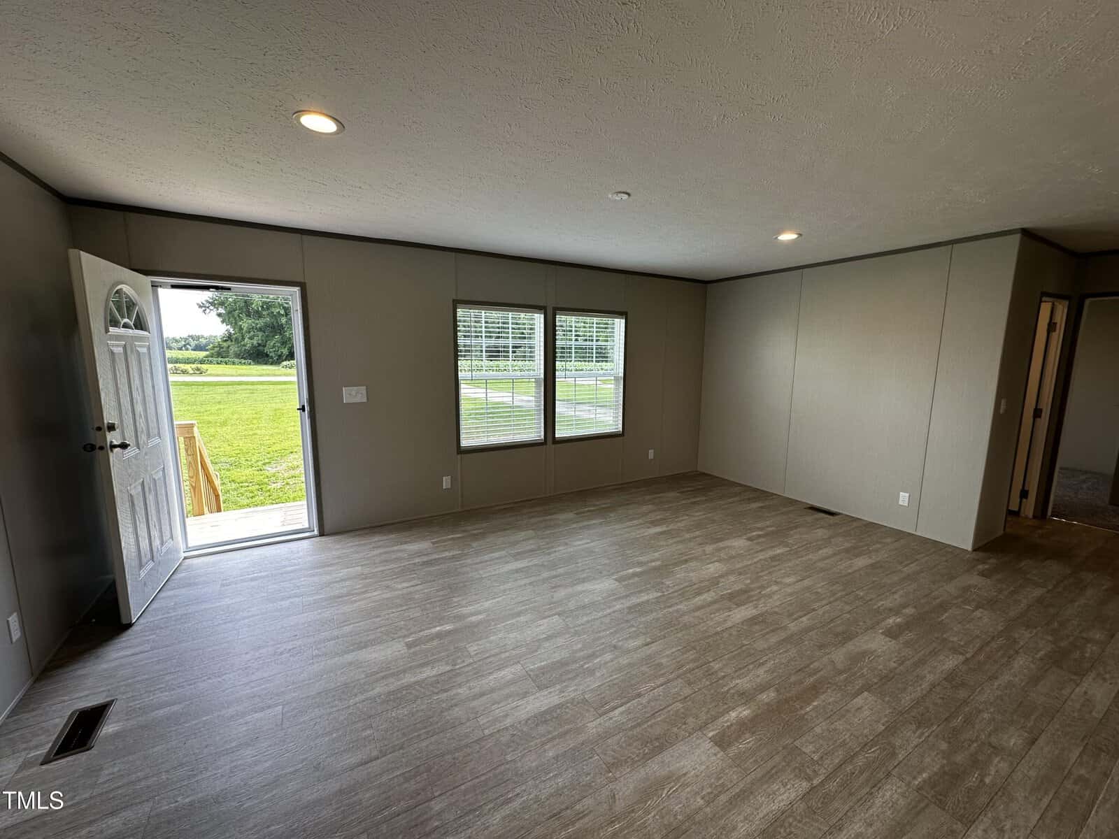 A spacious, empty room with wood-style flooring, beige walls, two large windows, and an open door leading outside to a grassy area with trees in the background. Ceiling lights are on, and natural light brightens the space.
