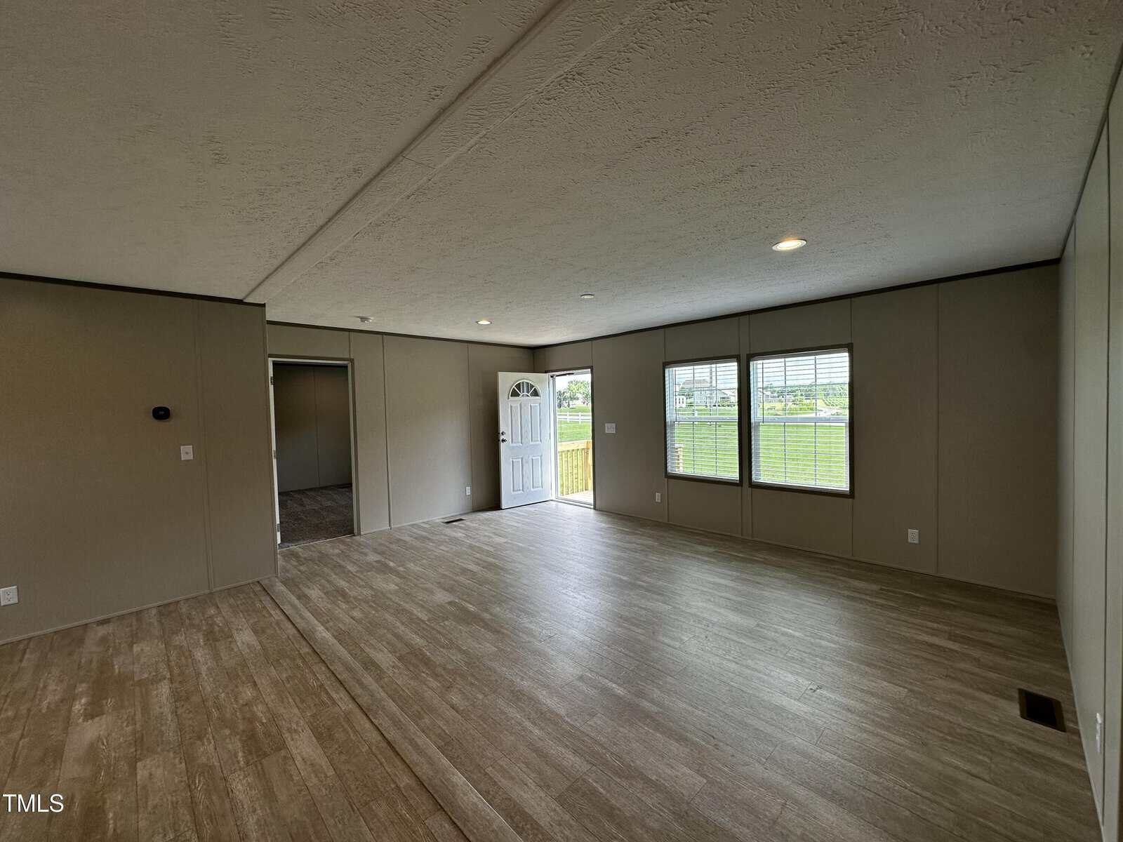 Spacious, empty living room with wood-style flooring, neutral walls, three large windows, and an open door revealing green grass outside. An adjacent room is partially visible through a doorway.