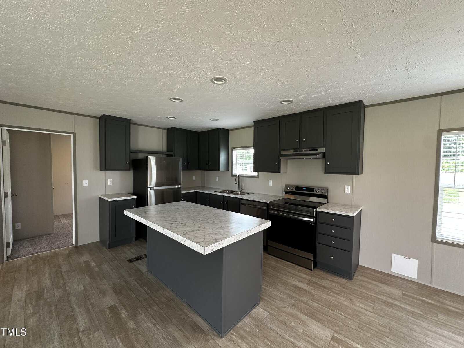 Modern kitchen with dark cabinets, stainless steel appliances, a marble-look island, and wood-style flooring. There are two windows, recessed ceiling lights, and a doorway leading to another room.
