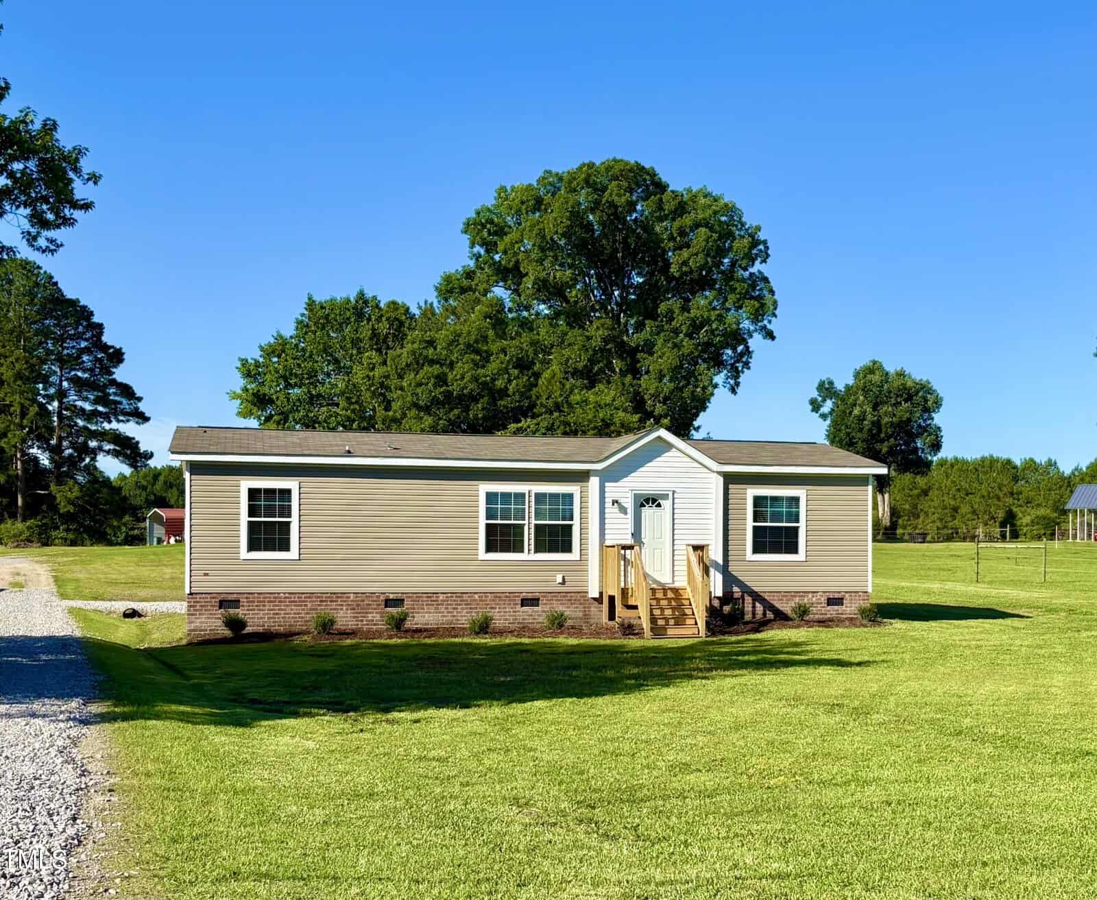 A single-story beige manufactured home with white trim and a small front porch, set on a large, well-manicured grassy lawn with trees and a gravel driveway under a clear blue sky.