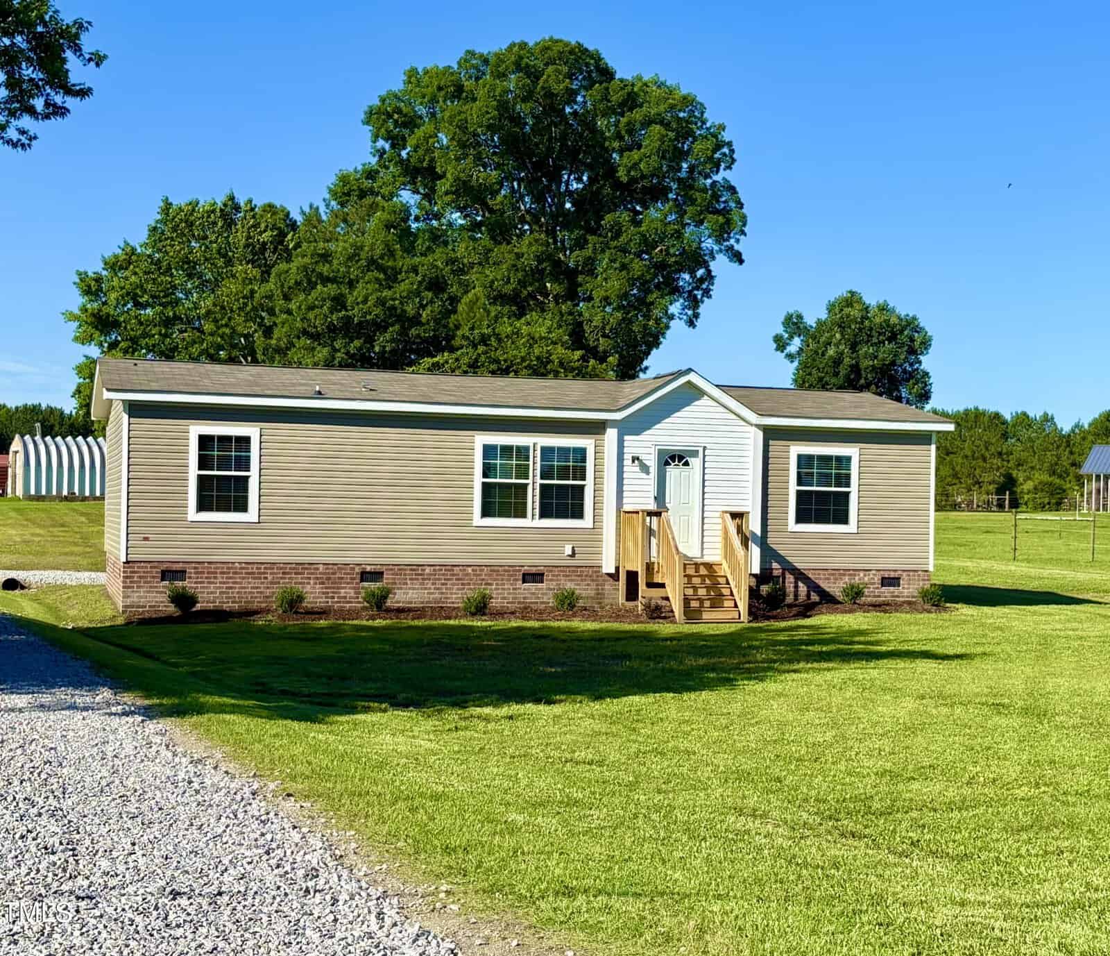 A beige manufactured home with white trim sits on a large green lawn, featuring a small front porch with steps, several windows, and brick skirting. Trees and a clear blue sky are in the background.