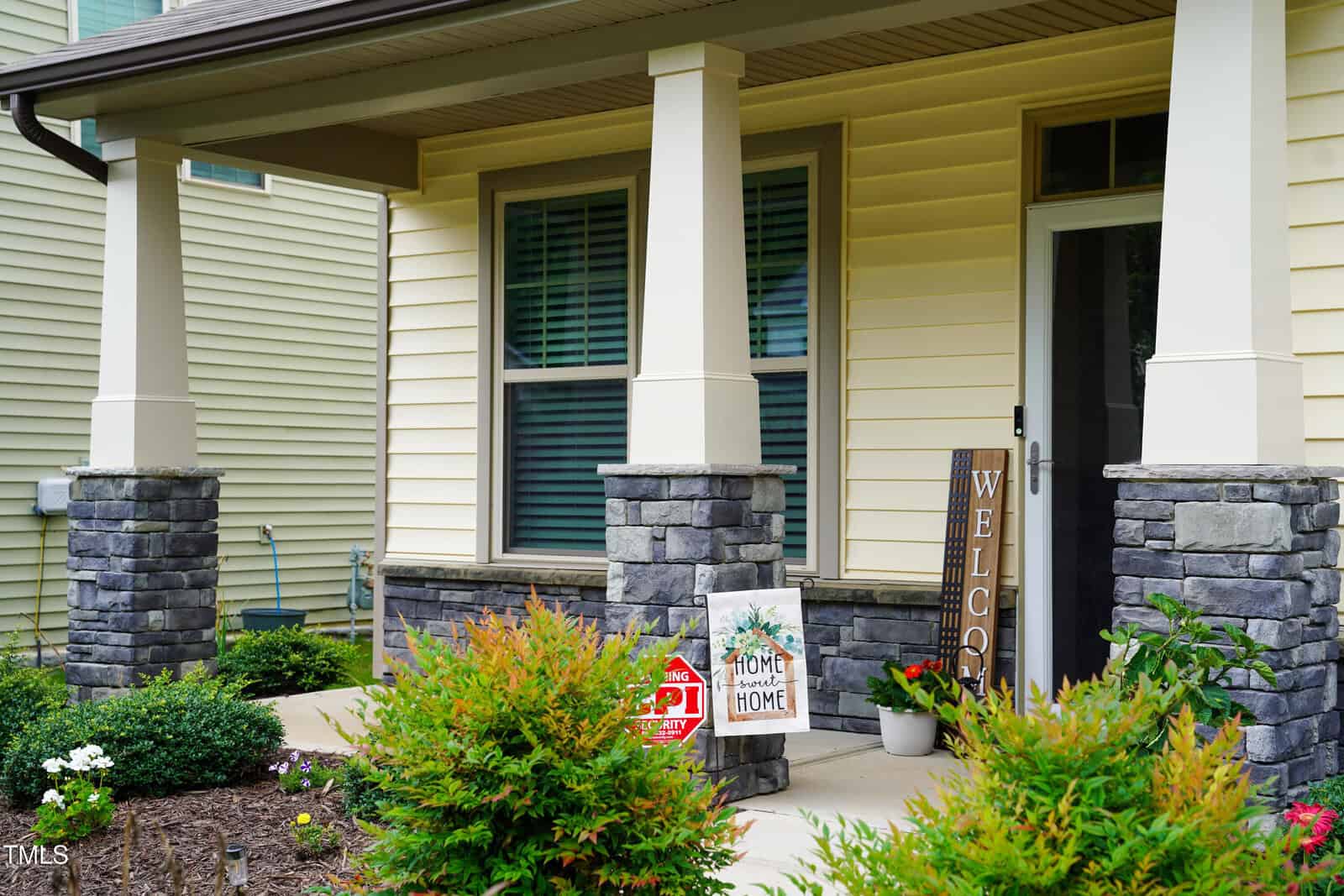 A house entryway with beige siding, stone pillars, a glass door, and welcome signs. There are potted plants and a small garden bed with shrubs in front of the porch. The area appears tidy and inviting.