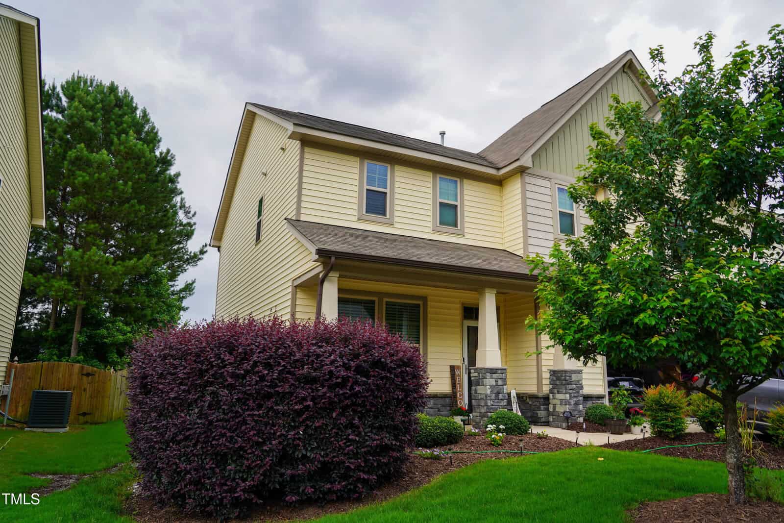 Two-story beige house with a covered front porch, stone accents, and white trim. There are bushes, a tree, and neatly maintained grass in the front yard. The sky appears cloudy.