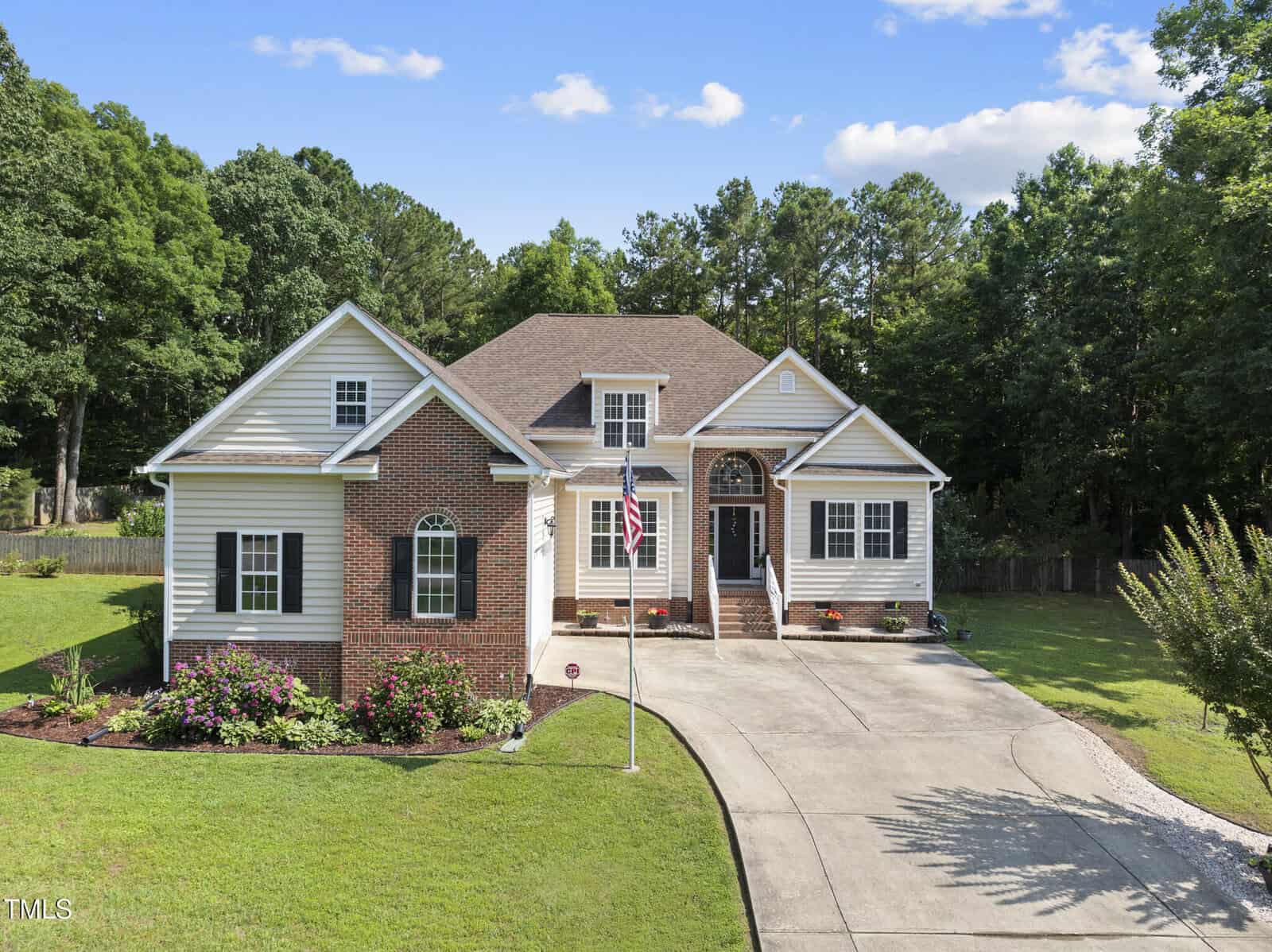 Single-story house with brick and siding exterior, arched entryway, and a gabled roof. A curved driveway leads to the front entrance, and the yard is landscaped with grass, shrubs, and flowers. Tall trees surround the property.