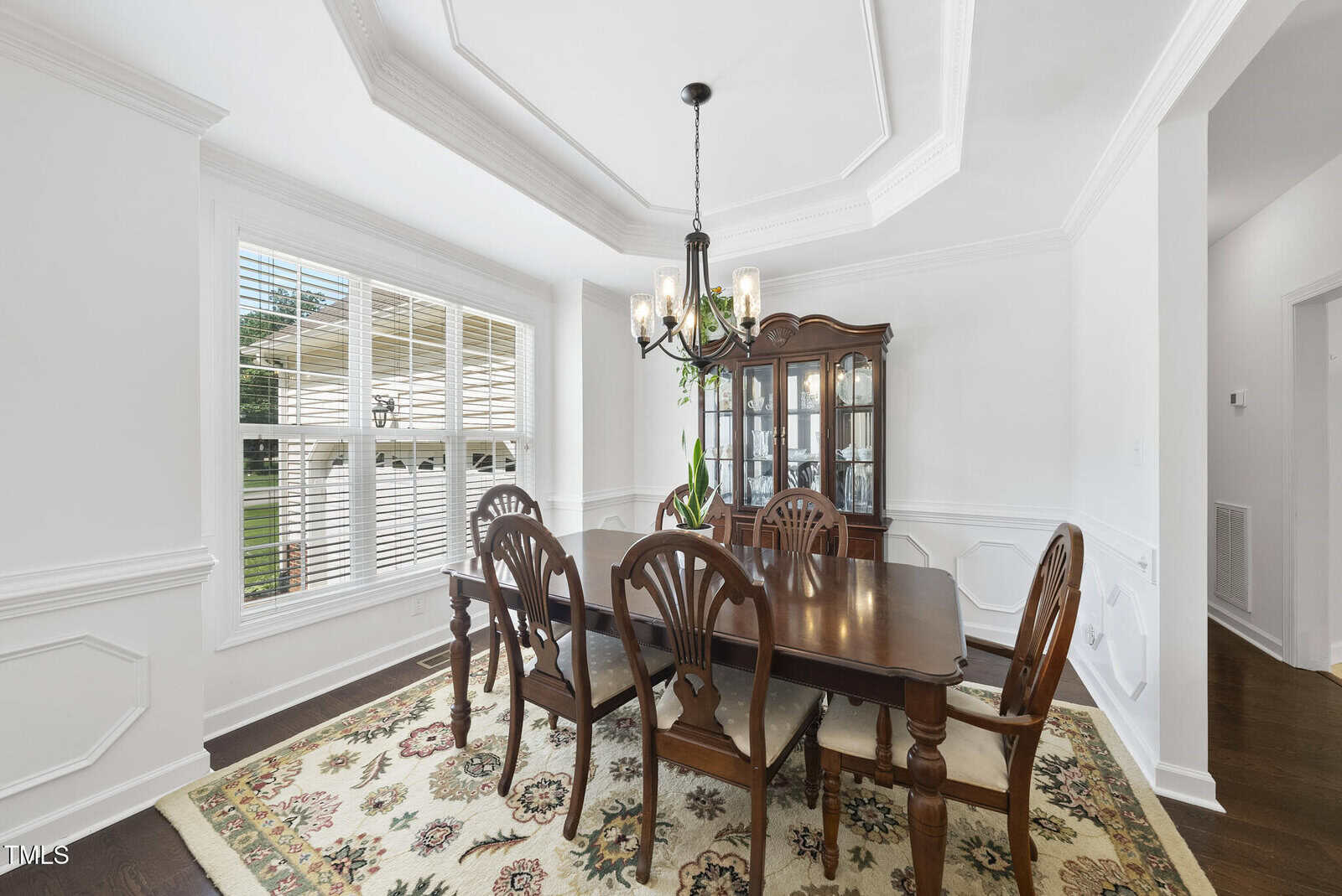 A dining room with a dark wooden table and six chairs, a glass-front china cabinet, a chandelier, large windows with blinds, white walls with wainscoting, and a patterned rug on a dark wood floor.