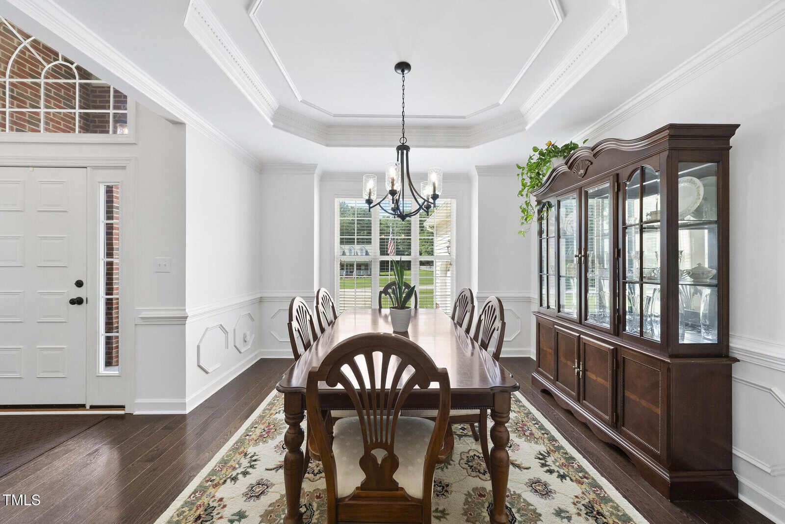 Bright dining room with a long wooden table, six chairs, a chandelier, a large window, a glass-front china cabinet, white walls, dark wood floors, and a floral rug. A plant sits on top of the cabinet.