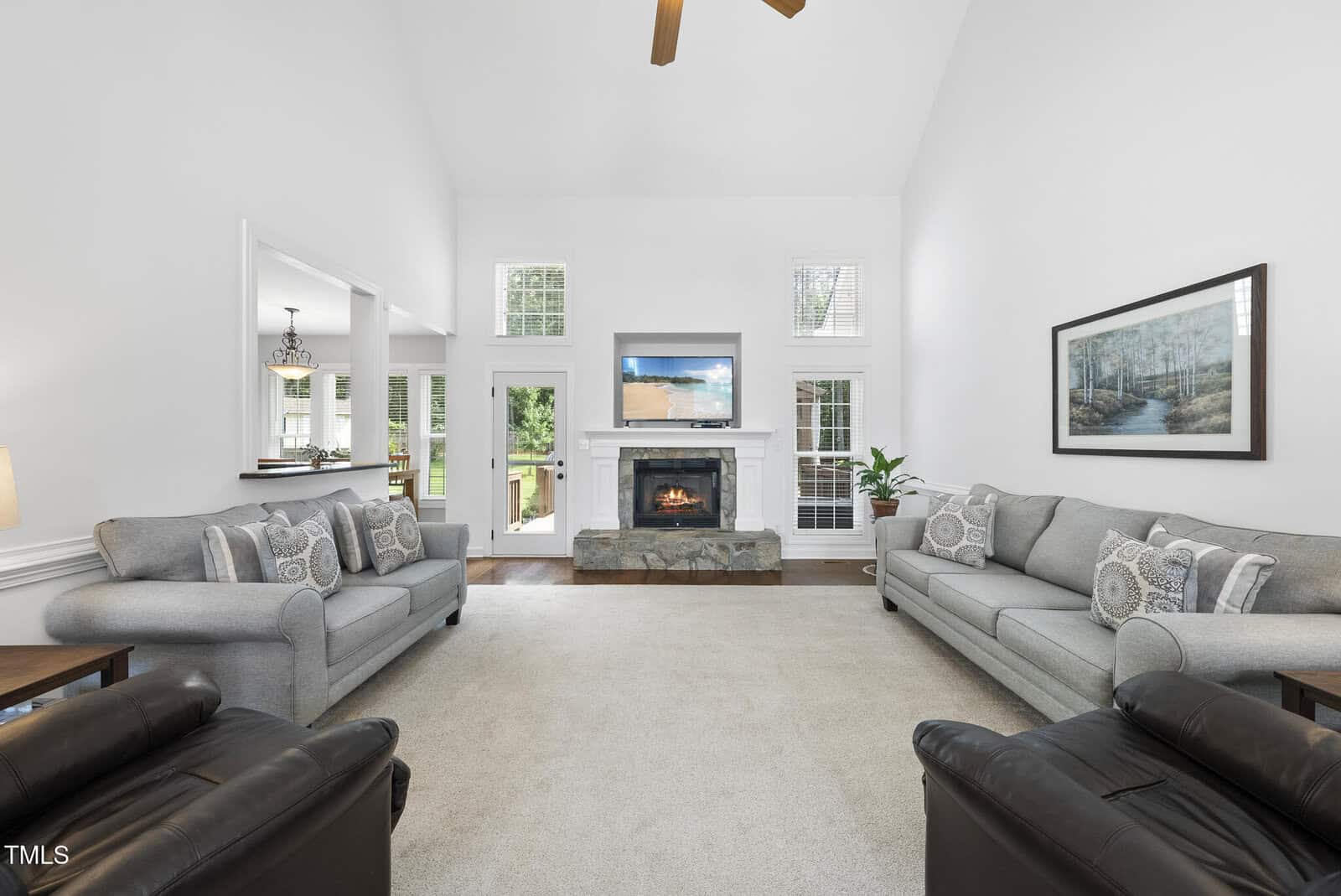 Bright living room with two gray sofas, two black armchairs, a stone fireplace, wall art, and a TV above the mantel. Large windows let in natural light, and an open doorway leads to a dining area.