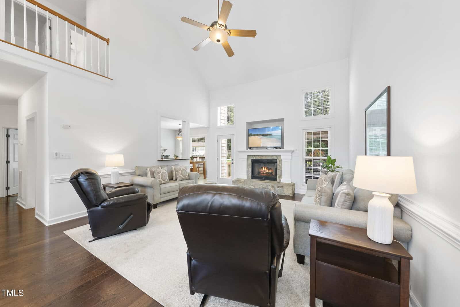 Bright living room with high ceiling, ceiling fan, fireplace, wall-mounted TV, two sofas, two leather recliners, rug, lamps, hardwood floors, and large windows letting in natural light. Kitchen visible through open doorway.