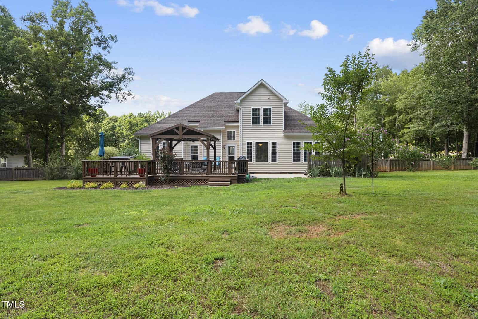 A two-story beige house with a spacious backyard, wooden deck, and gazebo. The yard is grassy with a few trees, and the scene has a clear blue sky with scattered clouds.