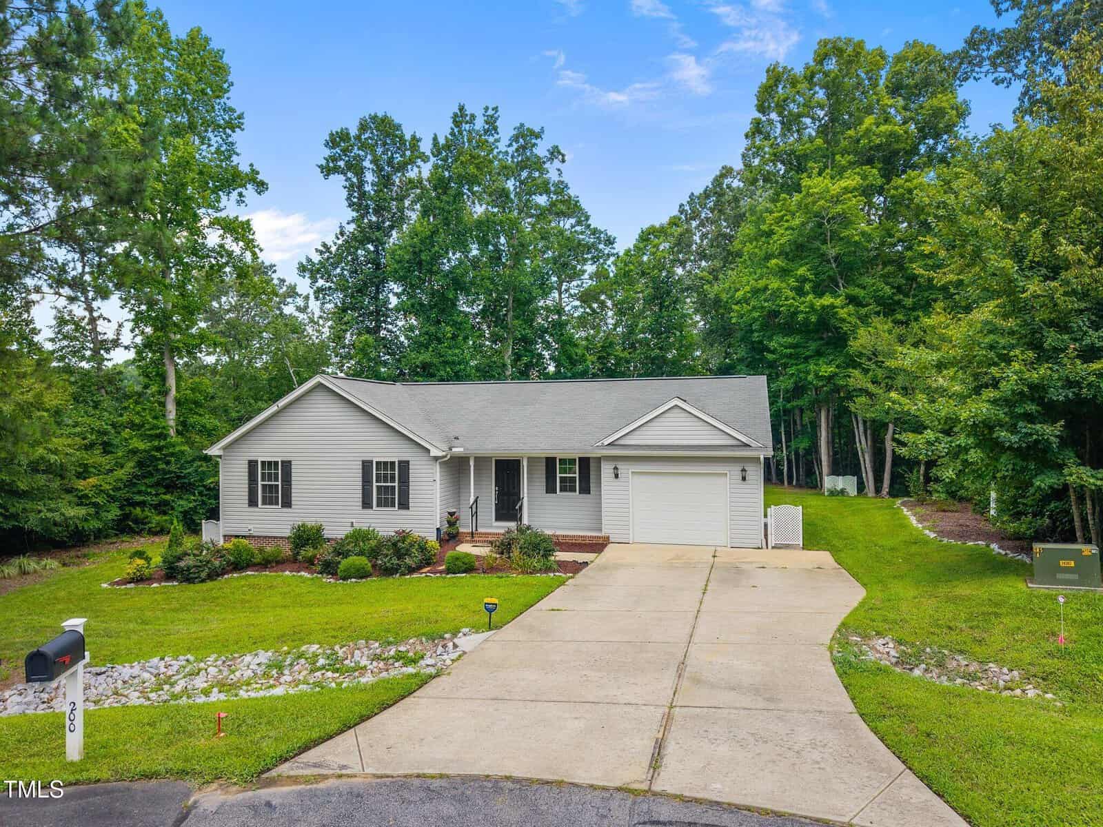 A single-story light gray house with a front porch, attached garage, wide driveway, and neatly landscaped yard, surrounded by tall green trees under a clear blue sky.