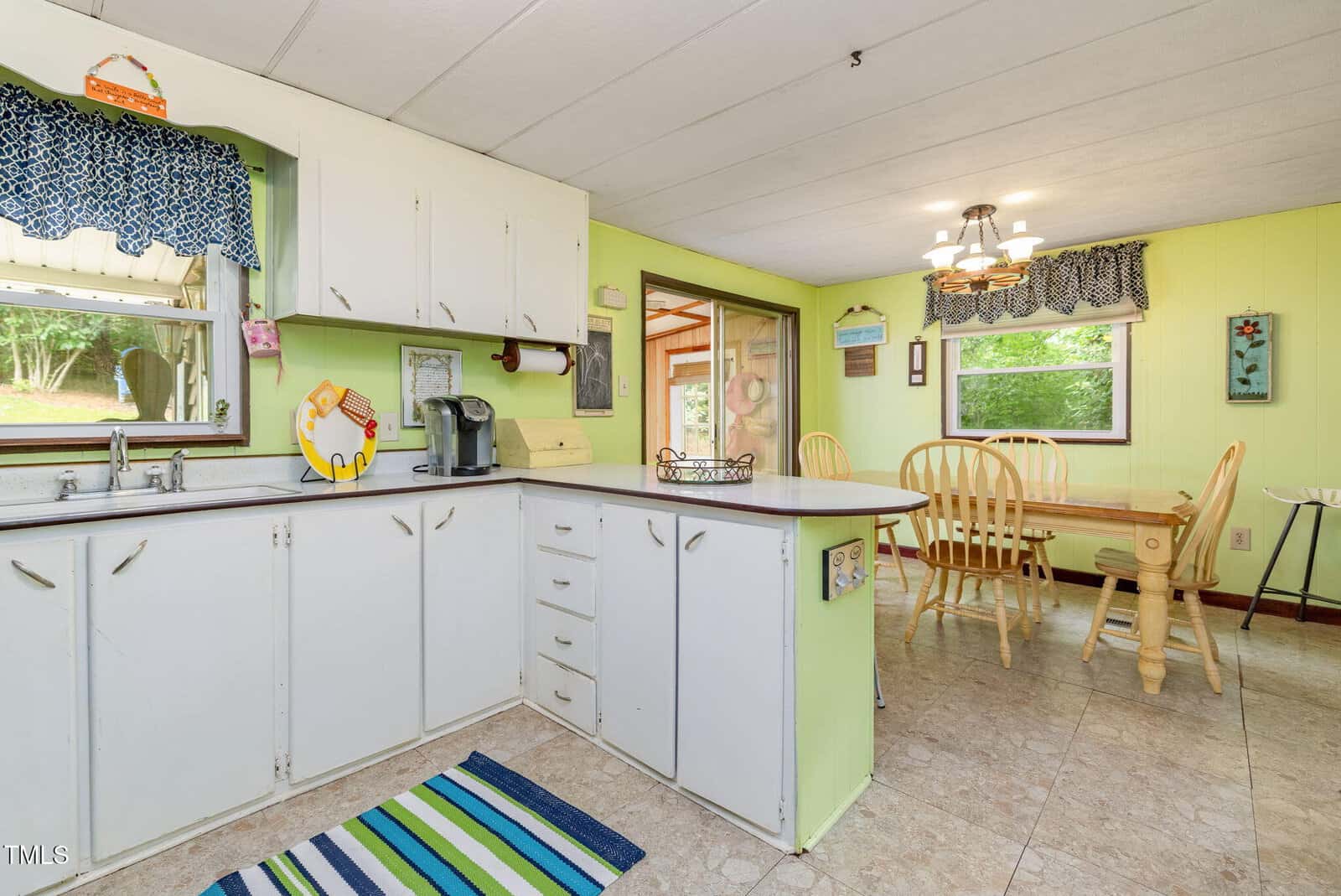 A kitchen with white cabinets, a coffee maker, and a striped rug leads into a dining area with a wooden table and chairs, green walls, and large windows letting in natural light.