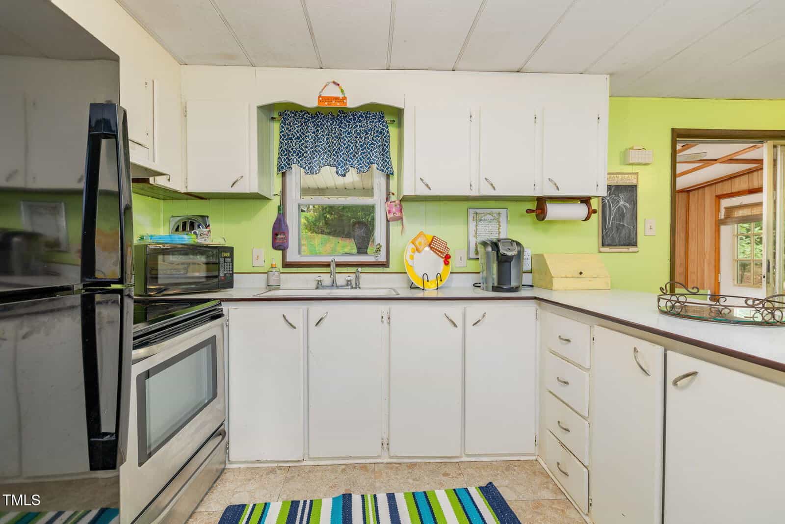 A kitchen with white cabinets and appliances, including a stove, microwave, and coffee maker. A window with a blue patterned valance is above the sink. The walls are painted light green. A colorful rug is on the floor.