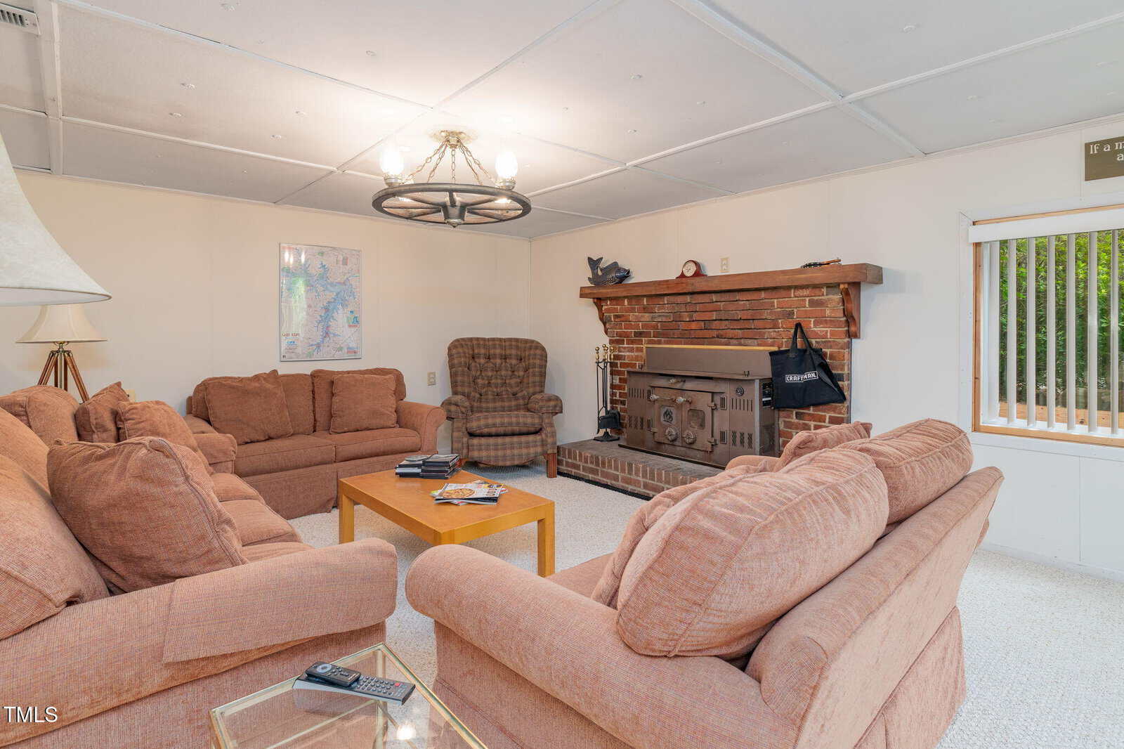 Cozy living room with peach sofas, wooden coffee table, armchair, brick fireplace with wood stove, map on the wall, and window with blinds, carpeted floor, and ceiling light fixture.