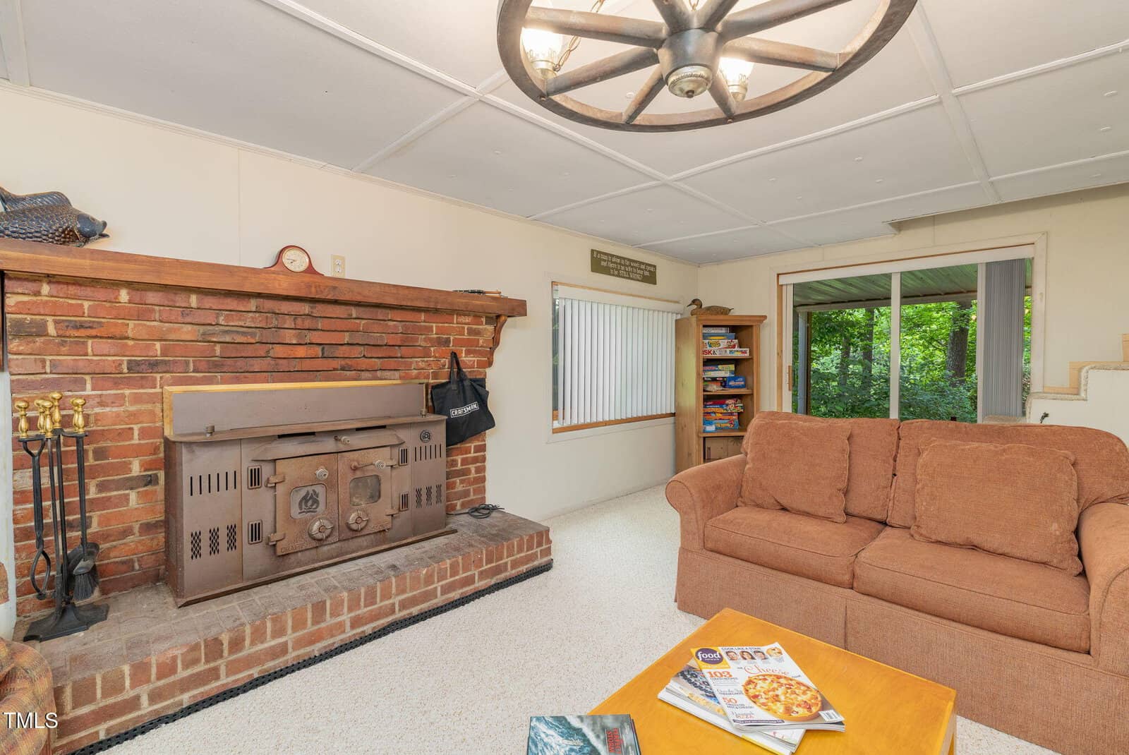 Cozy living room with a brown couch, wood-burning stove on a brick hearth, bookshelf, sliding glass door, and a wagon wheel ceiling light. A coffee table with magazines sits in the foreground.