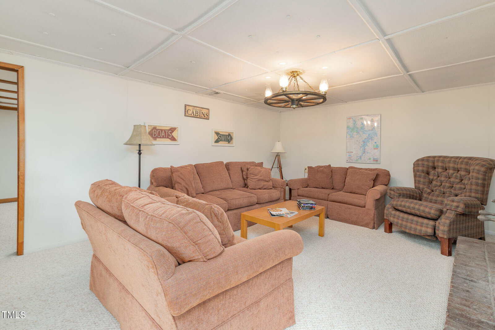 Cozy living room with two brown sofas, a plaid armchair, wooden coffee table, lamps, and wall decor including a map and rustic signs. Beige carpet and a chandelier complete the inviting space.