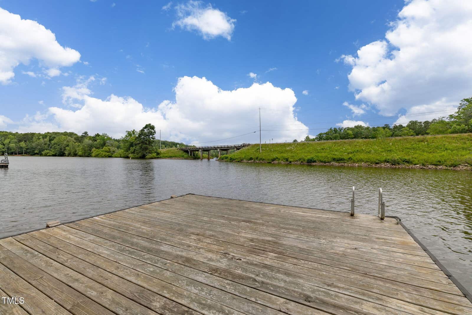 A wooden dock extends over a calm river with green grassy banks; a bridge and utility poles are visible in the background under a blue sky with scattered clouds.