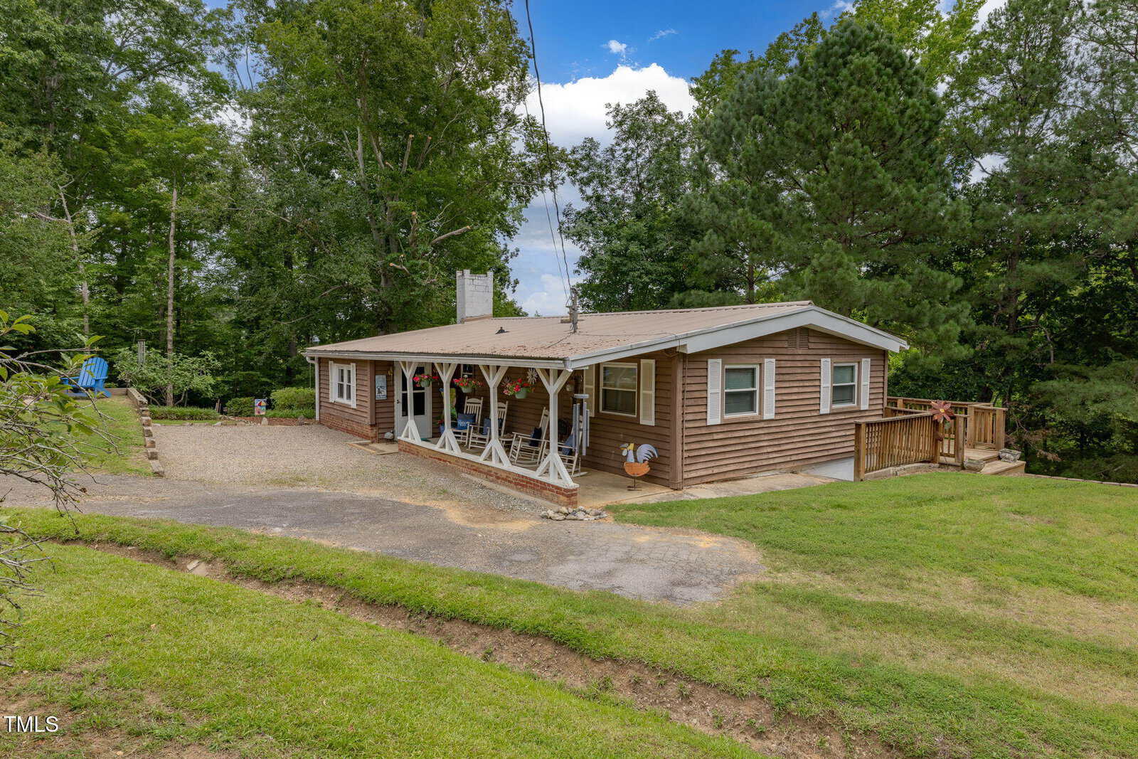 Charming single-story house at 394 Flanagan Road, Henderson, with brown siding, a covered front porch with decorative posts, paved driveway, lush green grass, trees, and outdoor seating under a partly cloudy sky.