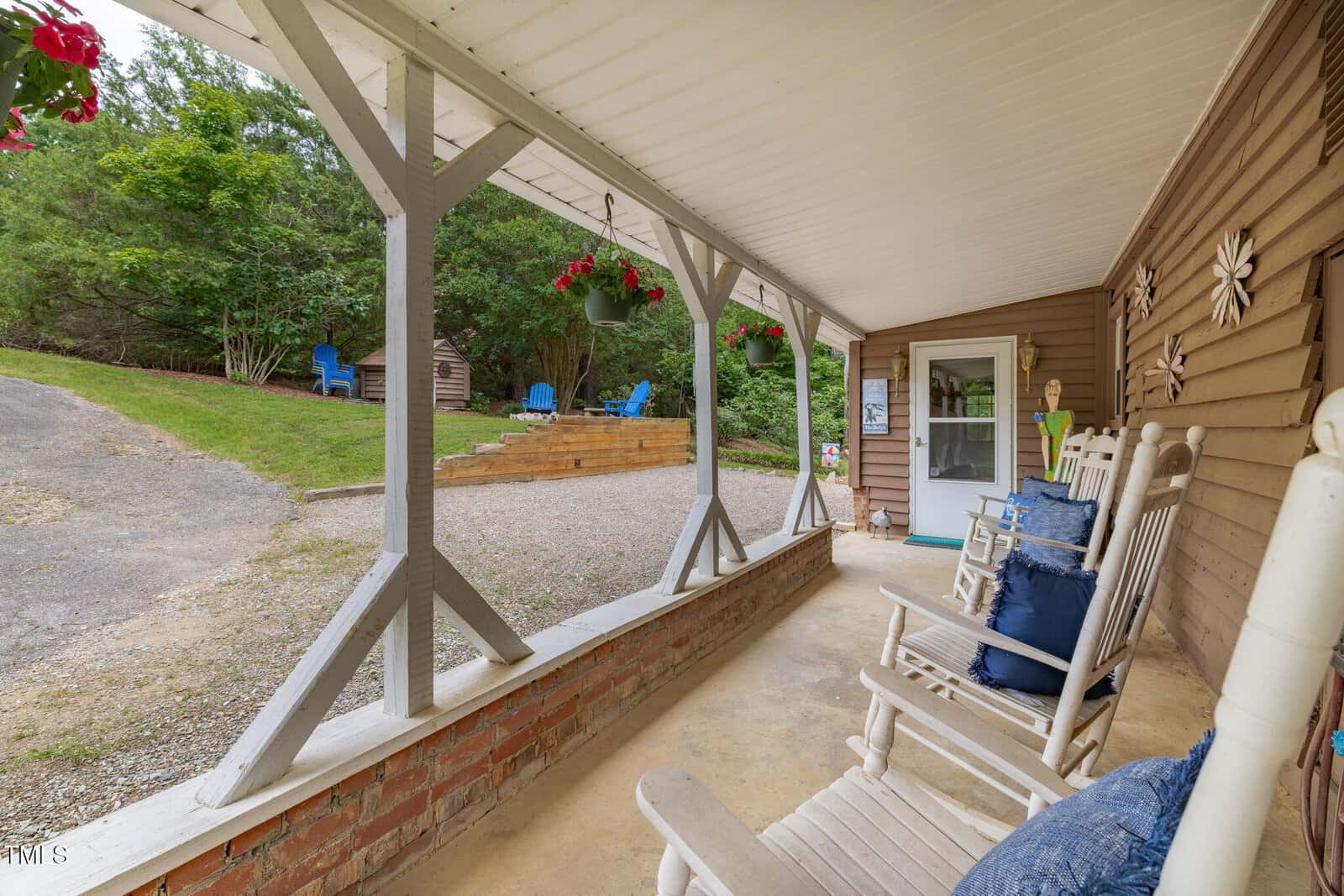 A covered porch with white rocking chairs and blue cushions faces a gravel driveway and a grassy area with Adirondack chairs. Hanging flower baskets and outdoor decorations are visible.