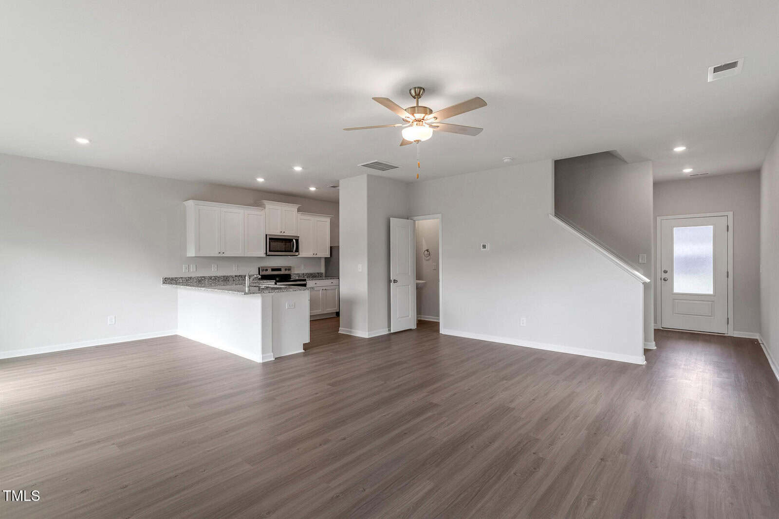 Unfurnished open-plan living area with light wood flooring, white walls, and ceiling fan; kitchen in the background with white cabinets and stainless steel appliances; staircase and back door visible.