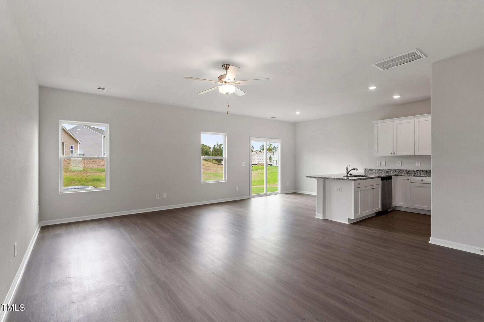 A spacious empty living area with light gray walls, dark wood flooring, a ceiling fan, and three windows. An open kitchen with white cabinets and granite countertops is visible in the corner. A door leads outside.