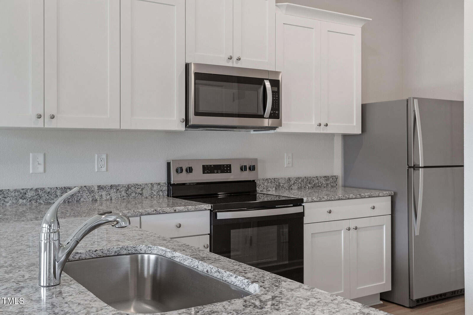 A modern kitchen features white cabinets, a granite countertop, a stainless steel oven with a black cooktop, a built-in microwave, a refrigerator, and a sink with a silver faucet.