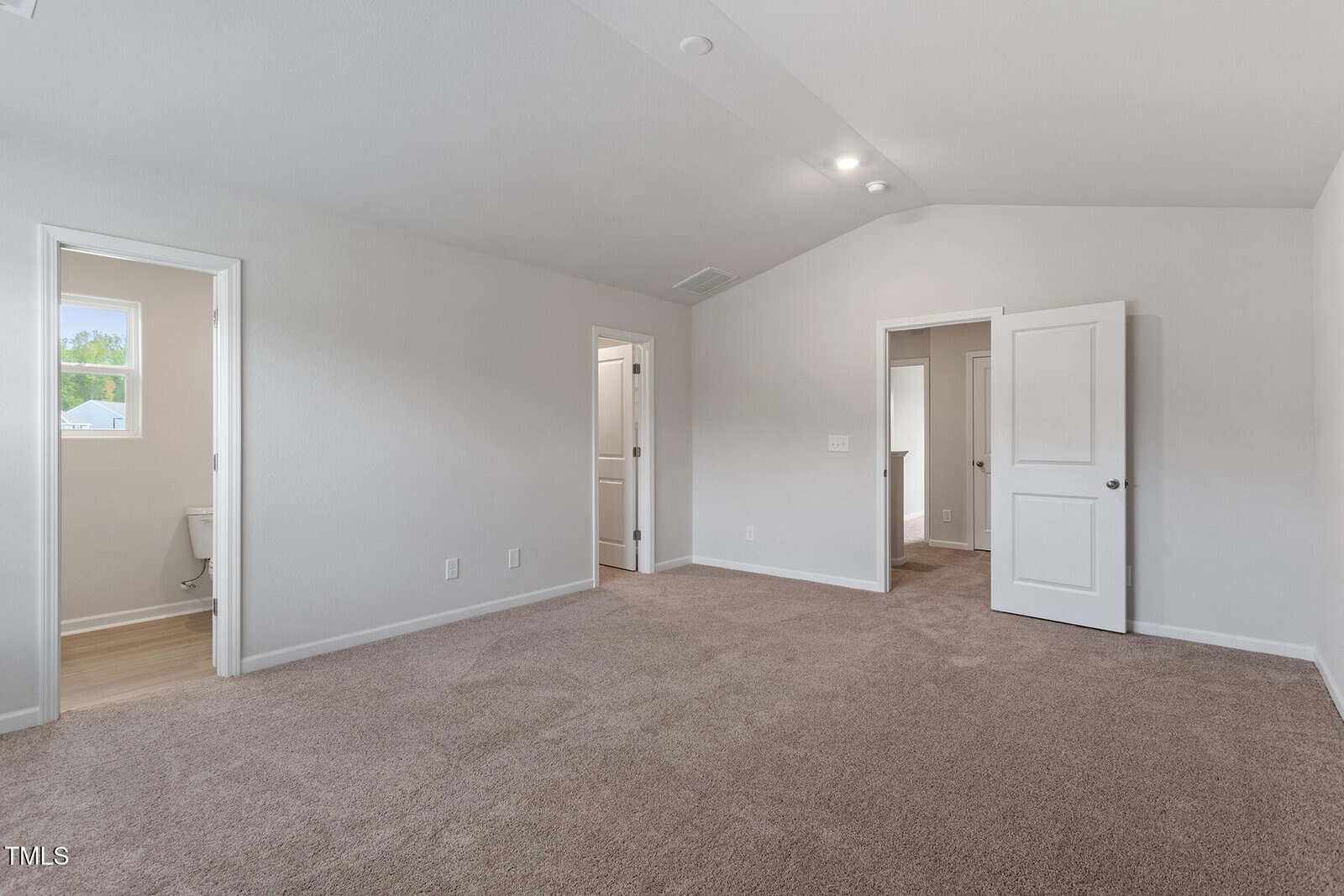 Empty carpeted bedroom with white walls, two open doors leading to a bathroom and closet, and a closed door on the right. Natural light enters through a small window near the bathroom. Ceiling lights are on.