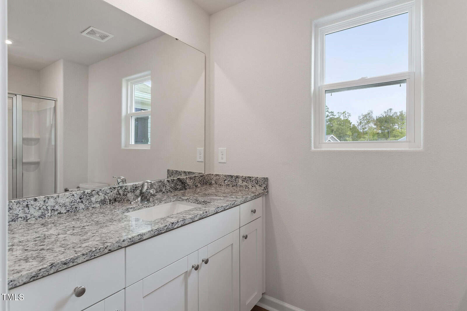 A modern bathroom with a white vanity, granite countertop, under-mount sink, two windows, and a partial view of a glass shower enclosure reflected in the large wall mirror.