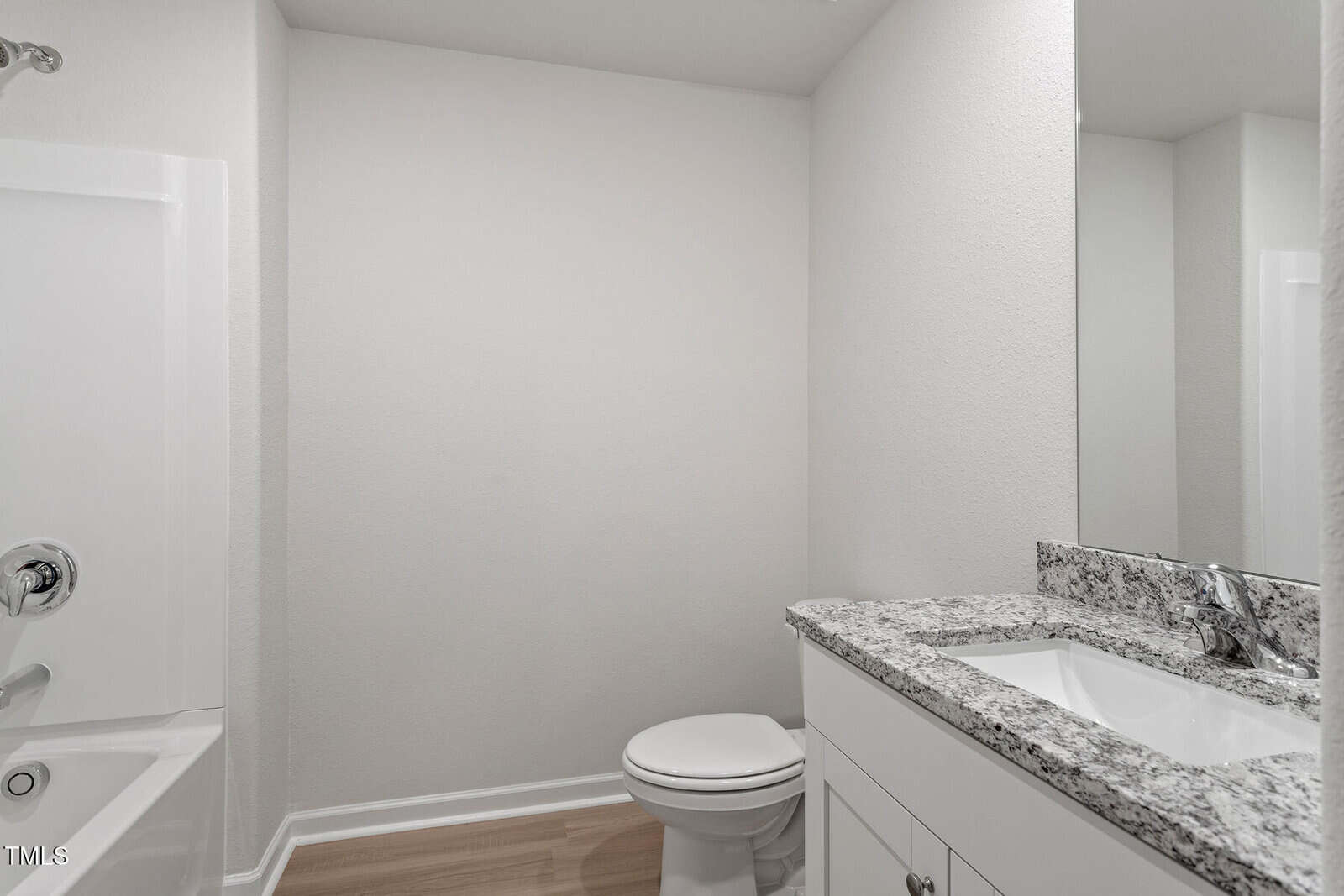 A modern bathroom with a white bathtub, toilet, and a granite countertop sink with cabinets beneath. The walls are light-colored, and the flooring is light wood. There is a large mirror above the sink.