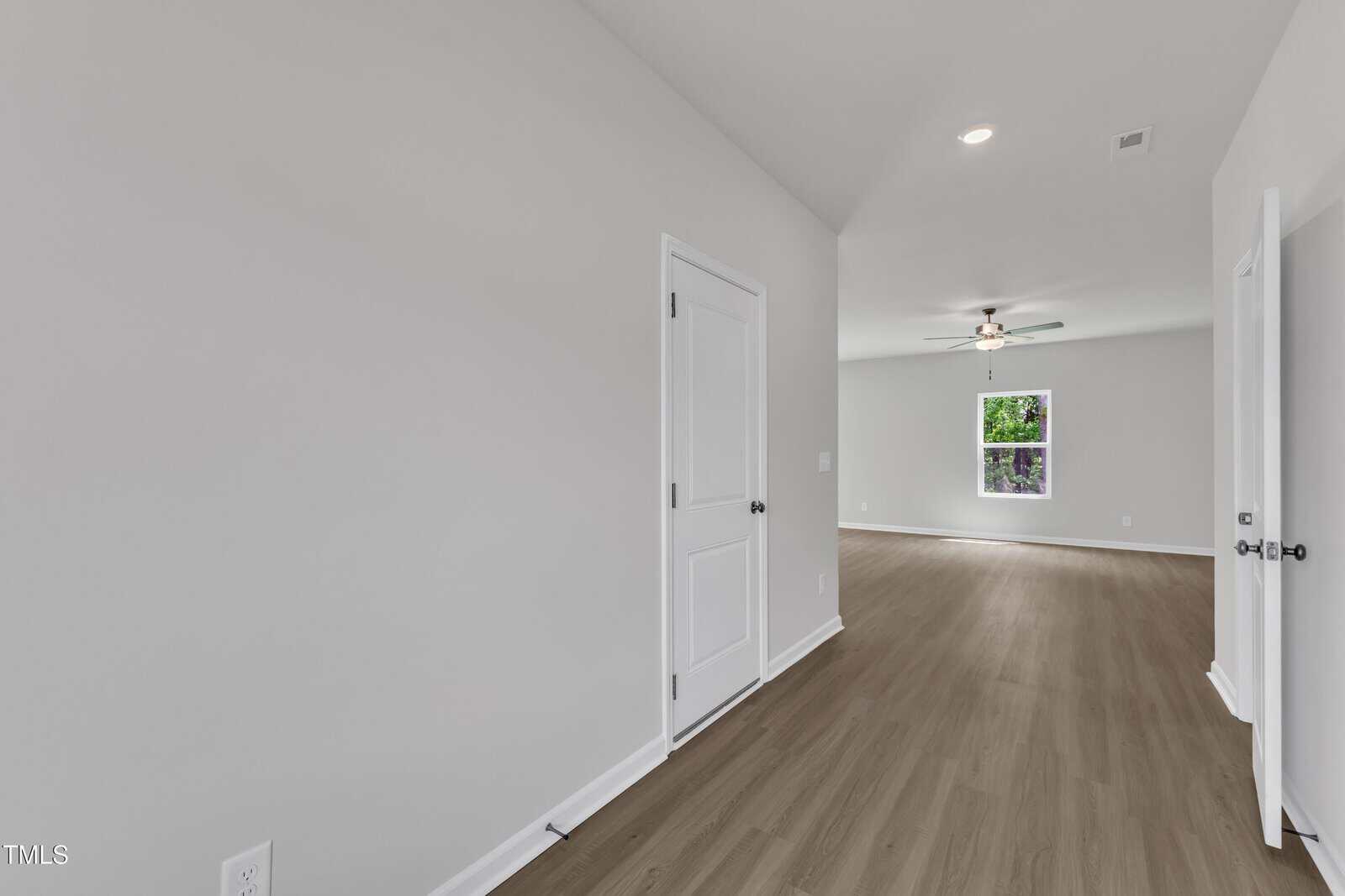 A view of an empty room with light-colored walls, wood flooring, two closed white doors on the left and right, a window at the far end, and a ceiling fan with light in the background.