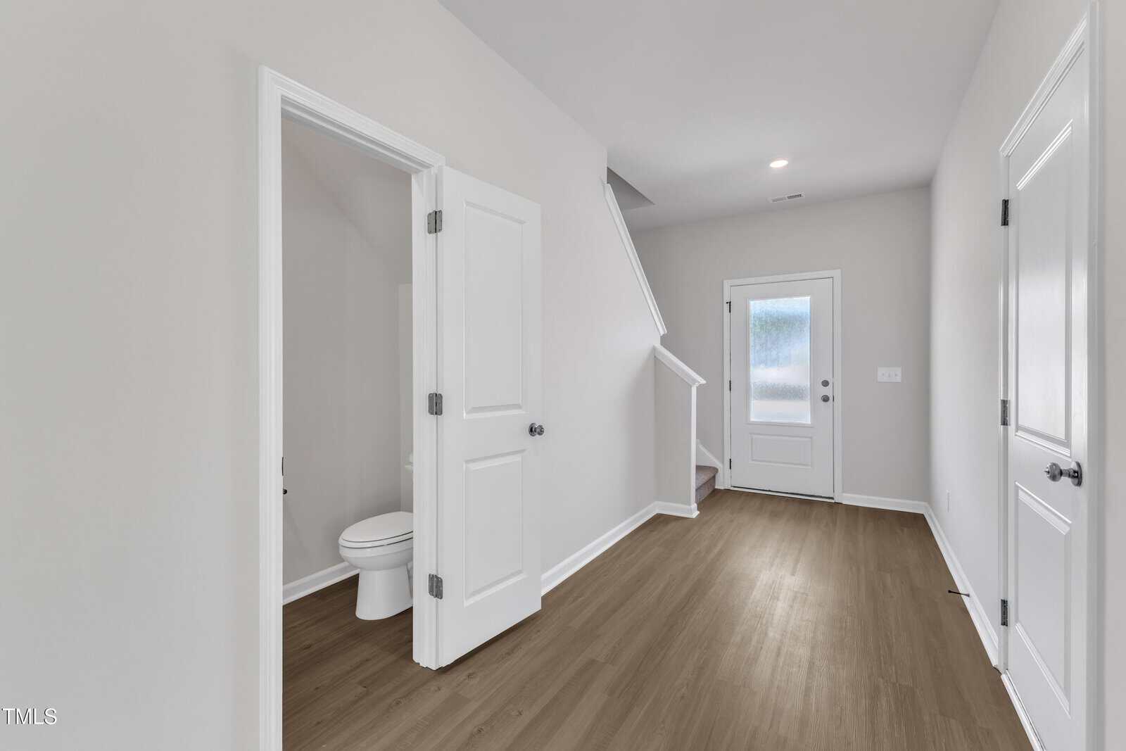 A hallway with light gray walls and wood flooring features a white door leading to a small bathroom with a toilet, a staircase, a back door with frosted glass, and another closed door on the right.