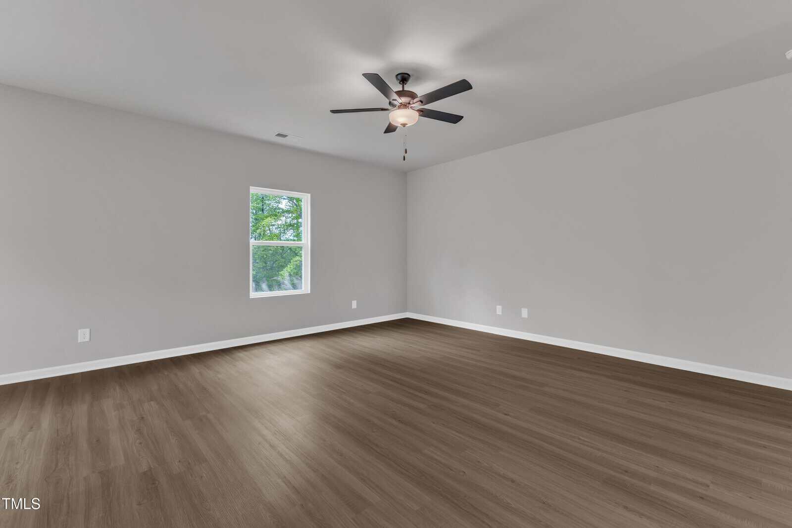 Empty room with light gray walls, a single window showing greenery outside, a ceiling fan with light, and wood-look flooring. Several electrical outlets are visible on the walls.