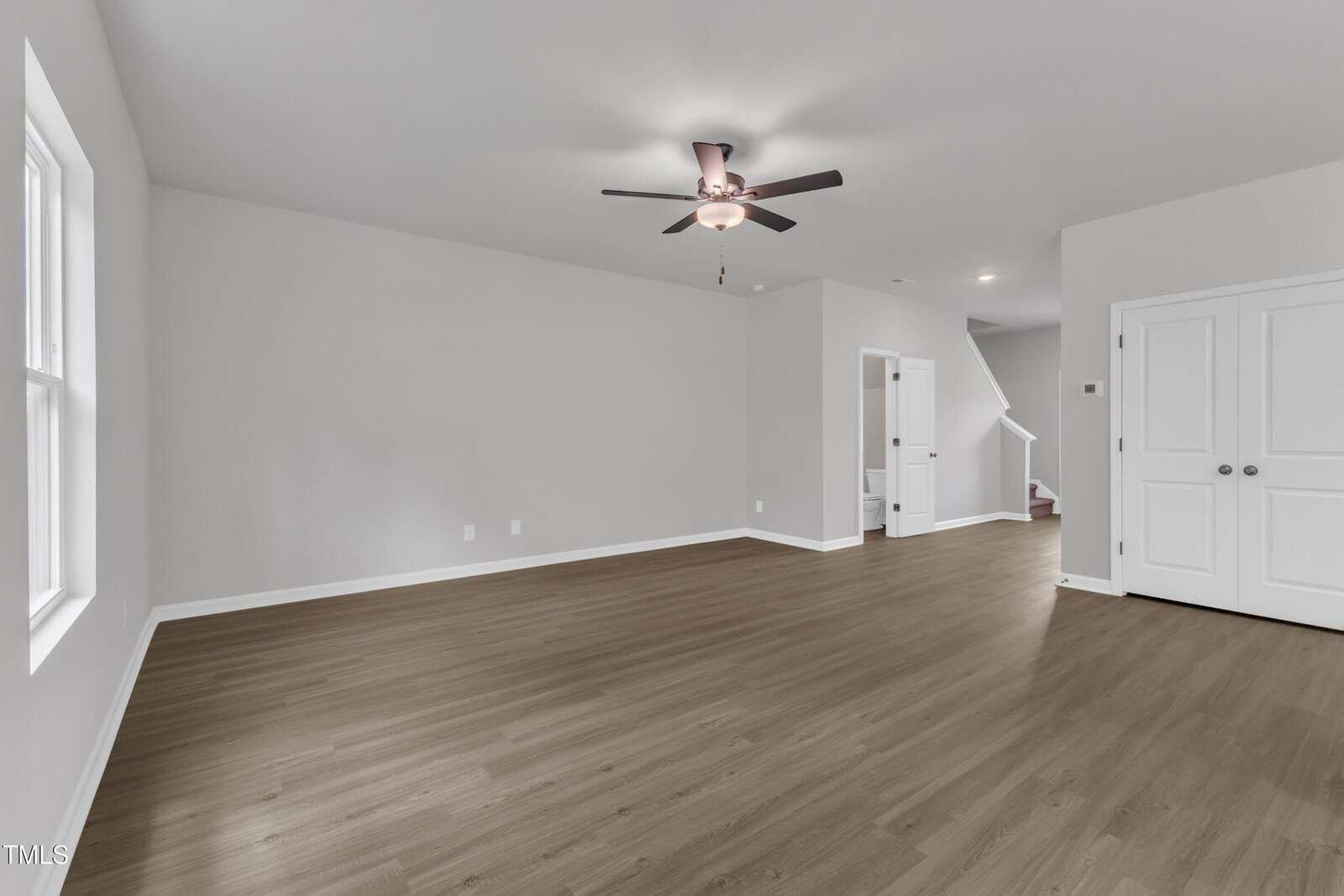 Spacious, empty room with light gray walls, wood-style flooring, a ceiling fan with lights, double doors on the right, and a staircase leading up at the back. Natural light enters from large windows on the left.
