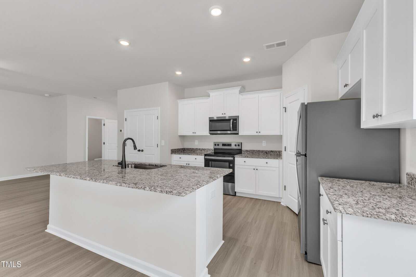 Modern kitchen with white cabinets, stainless steel appliances, granite countertops, a large central island with a black faucet, light wood flooring, and recessed ceiling lights. Walls are painted a light neutral color.
