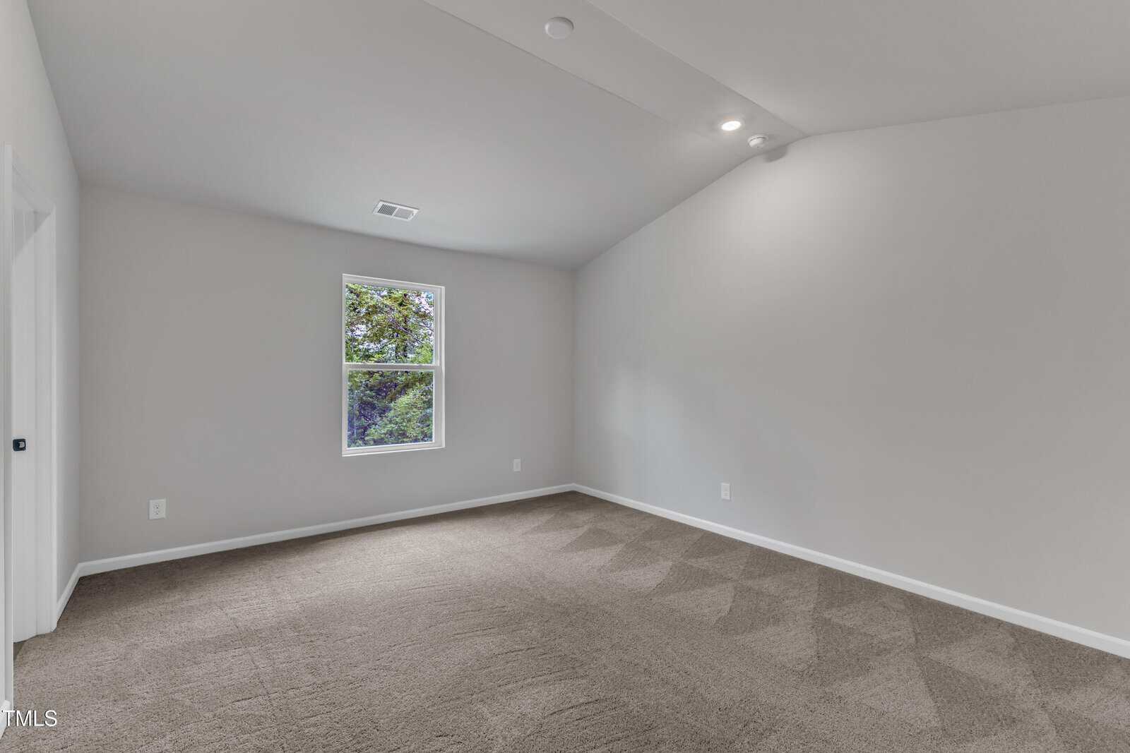 Empty room with light gray walls, a single window showing green trees outside, sloped ceiling, and clean brown carpet with visible vacuum lines. The room is well-lit with natural and ceiling lights.