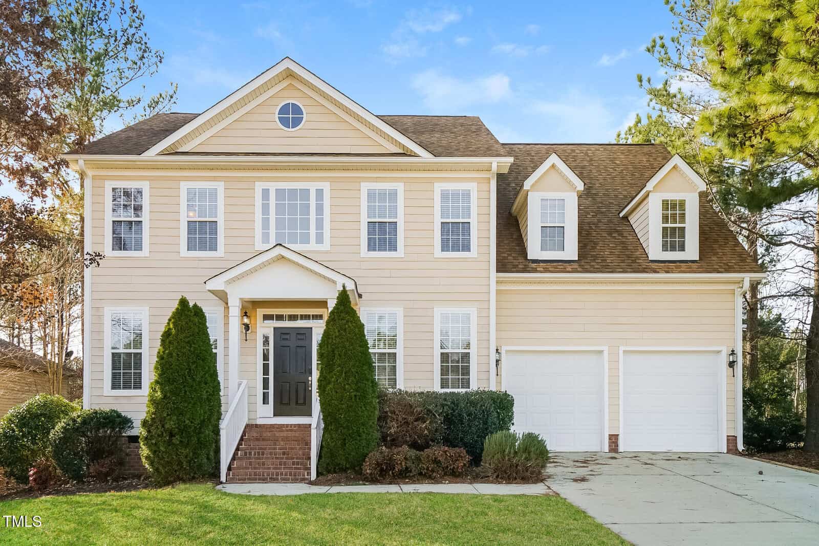 A two-story beige house with white trim, black front door, and two-car garage. Tall shrubs and small bushes line the front, with a paved driveway and steps leading to the entrance. Pine trees and blue sky are in the background.