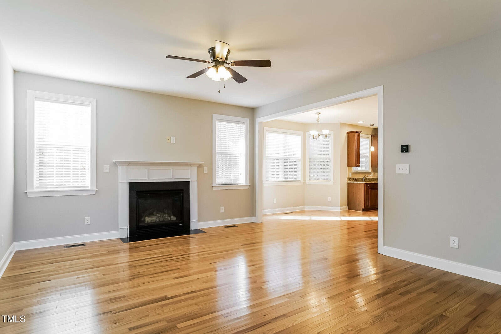 Bright living room with hardwood floors, a ceiling fan, white fireplace, and large windows. An open doorway leads to a kitchen and dining area with tile flooring and wooden cabinets.