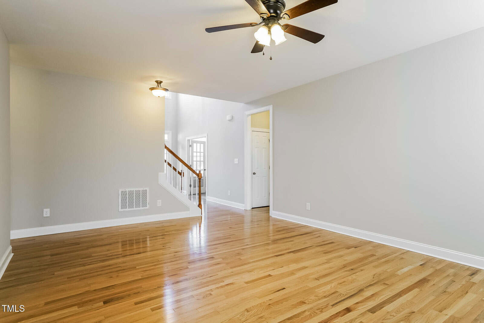 Bright, empty living room with light hardwood floors, beige walls, white baseboards, ceiling fan with lights, and a view of a staircase and white door in the background.
