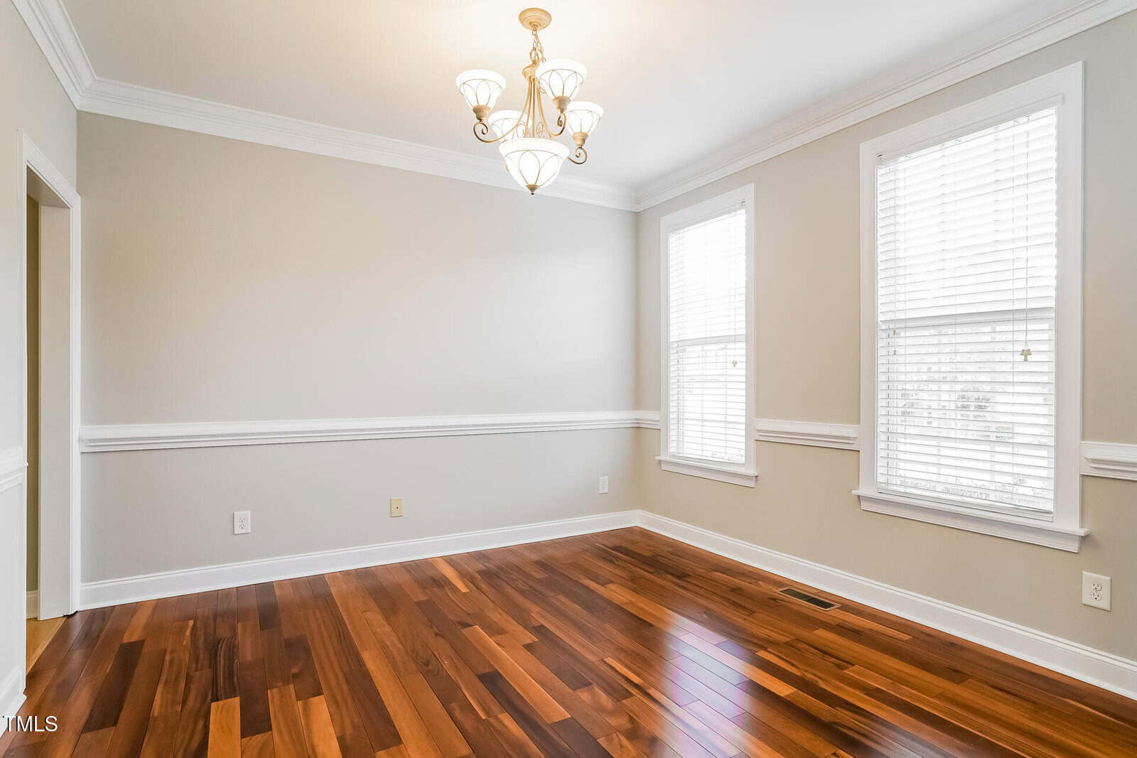 A bright, empty room with light gray walls, white trim, two large windows with blinds, a central ceiling light fixture, and polished hardwood flooring.