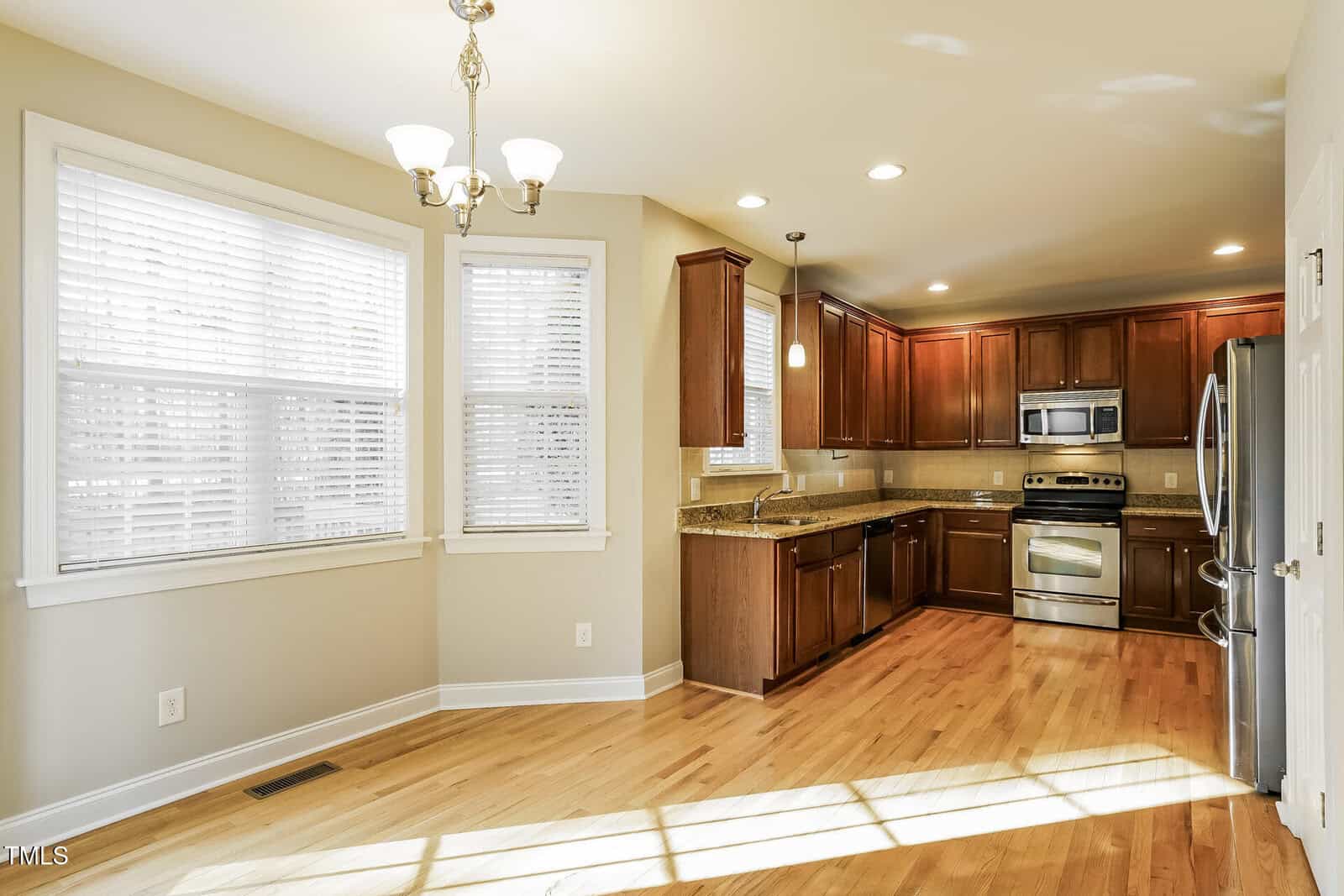 Bright kitchen with stainless steel appliances, dark wood cabinets, granite countertops, light hardwood floors, and a dining nook with large windows and white blinds.