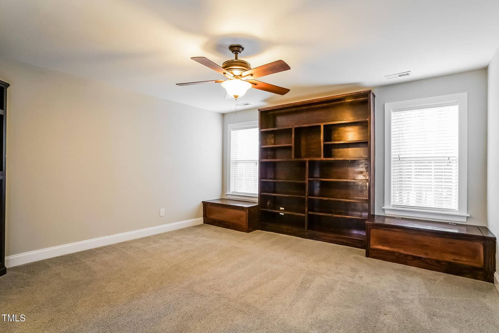 Empty carpeted room with neutral walls, a ceiling fan with light, two windows with blinds, and a large dark wood built-in bookcase flanked by two window seats with storage. The room is well-lit by natural light.