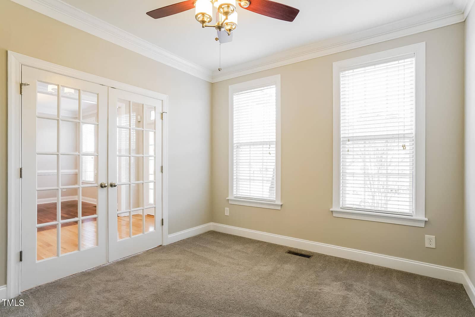 A small room with beige walls, two large windows with blinds, carpeted floor, a ceiling fan with lights, and double glass French doors leading to a room with hardwood flooring.